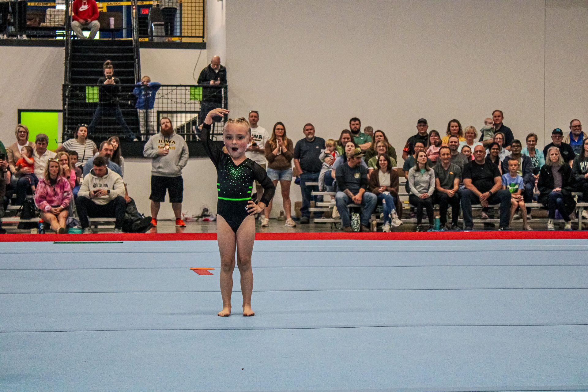 Gymnast in a black leotard standing on a blue floor mat during a competition, with spectators seated behind her