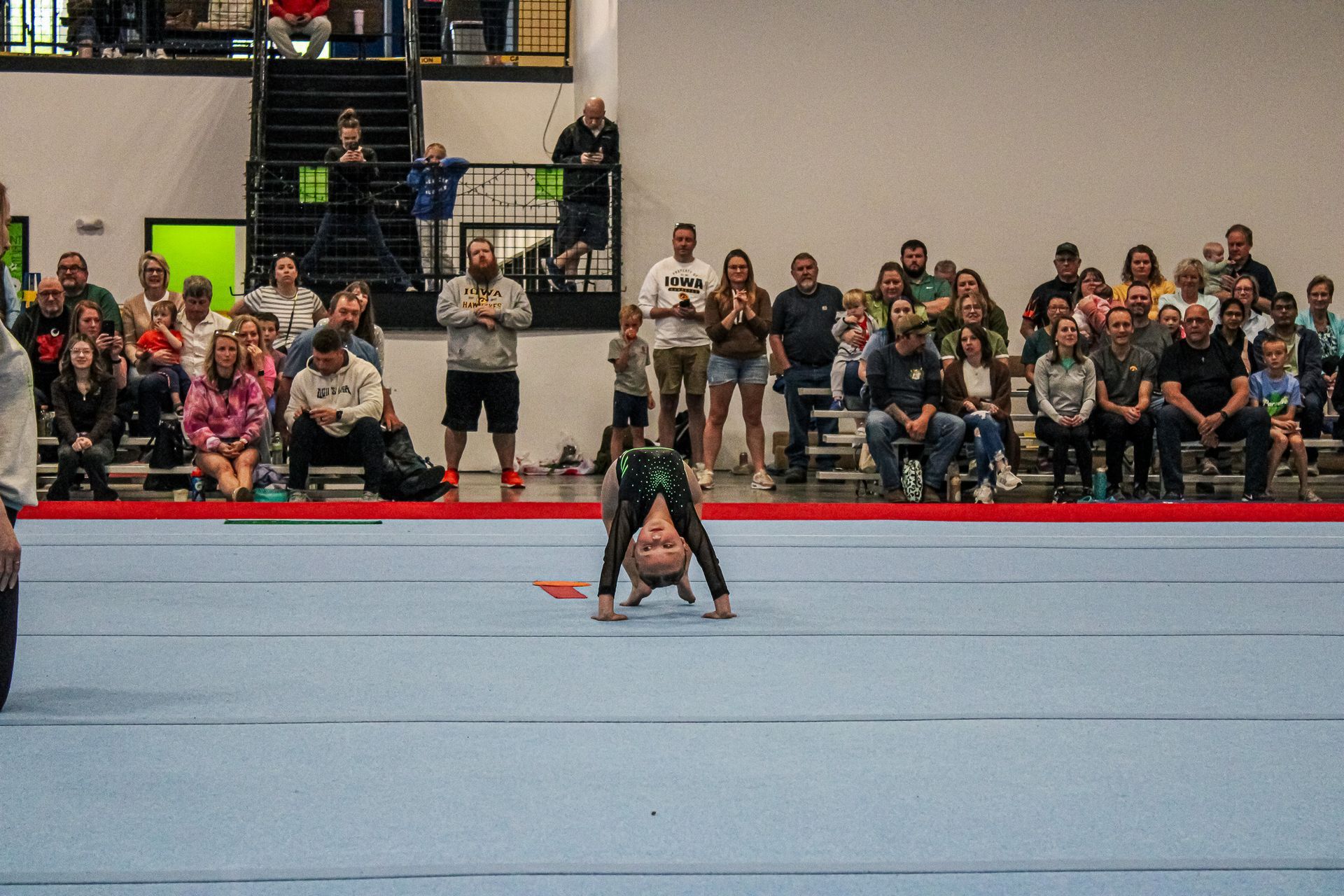 Gymnast crouched on blue mat in arena, with seated spectators in the background