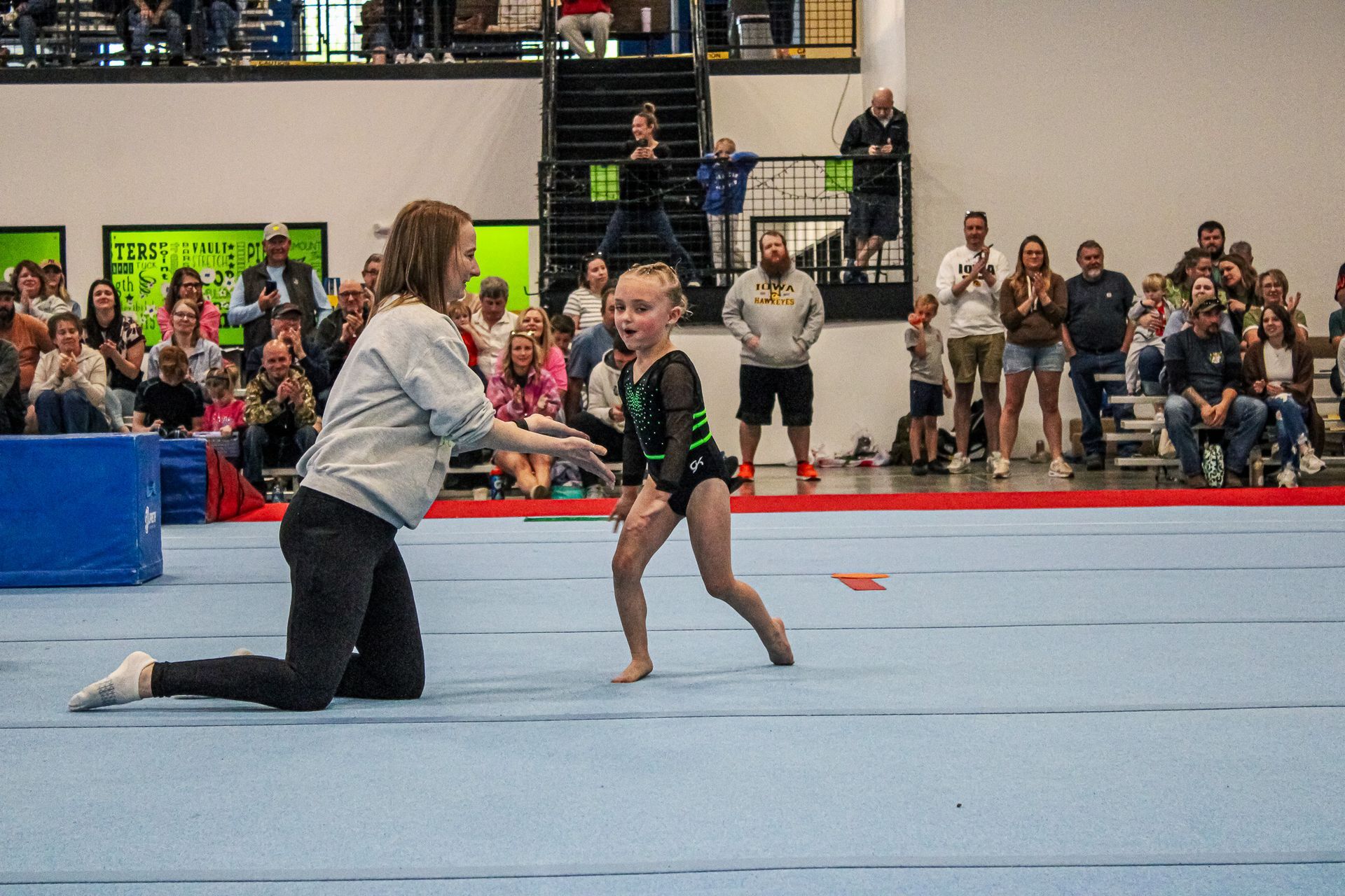 Gymnast kneeling on the mat guiding a young gymnast in a gym with spectators in the background