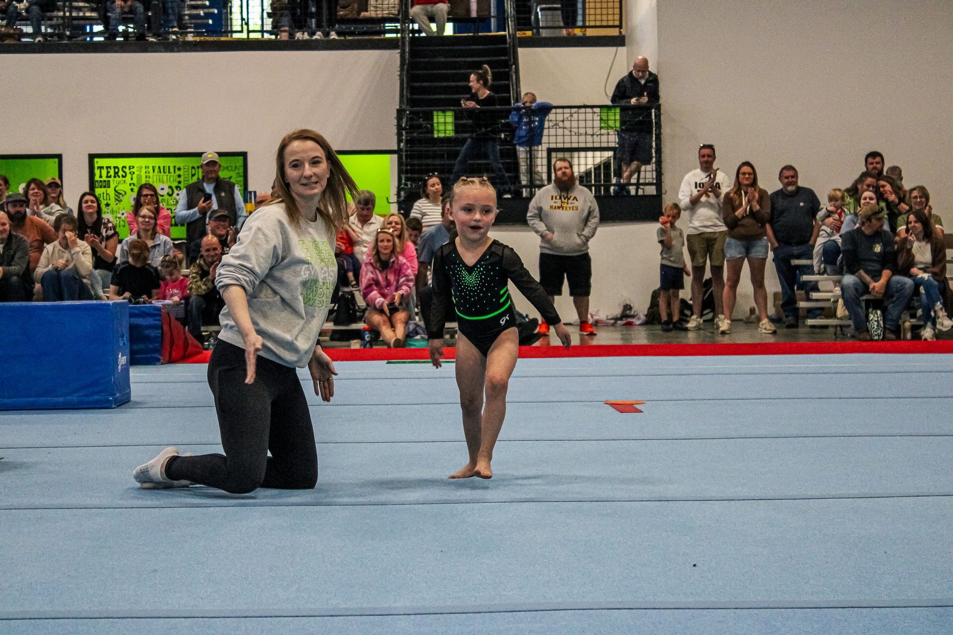 Gymnast and coach on blue floor mat during a performance, with spectators watching in the background.