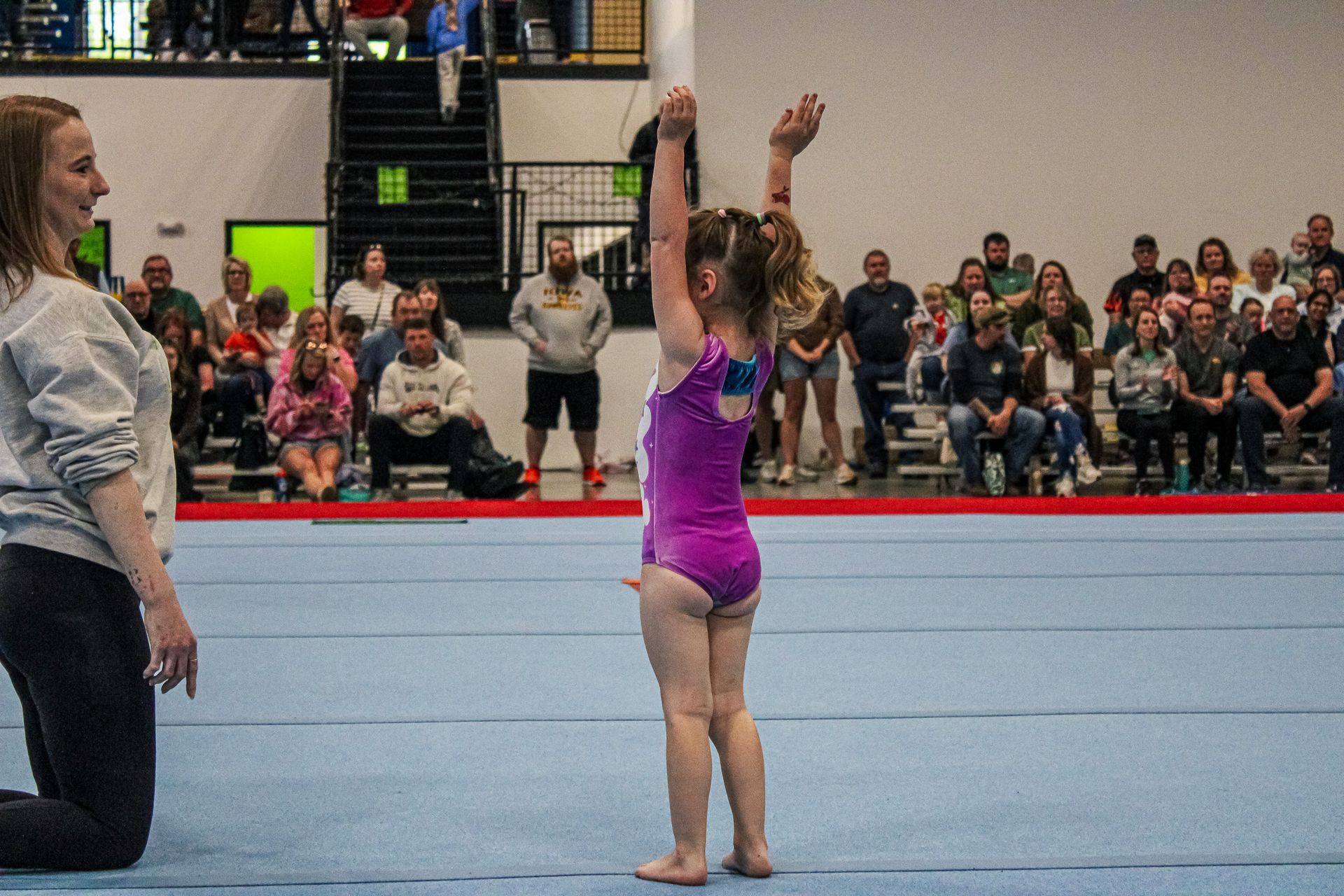 Gymnast in purple leotard raises arms on a mat, with a coach and seated audience in the background