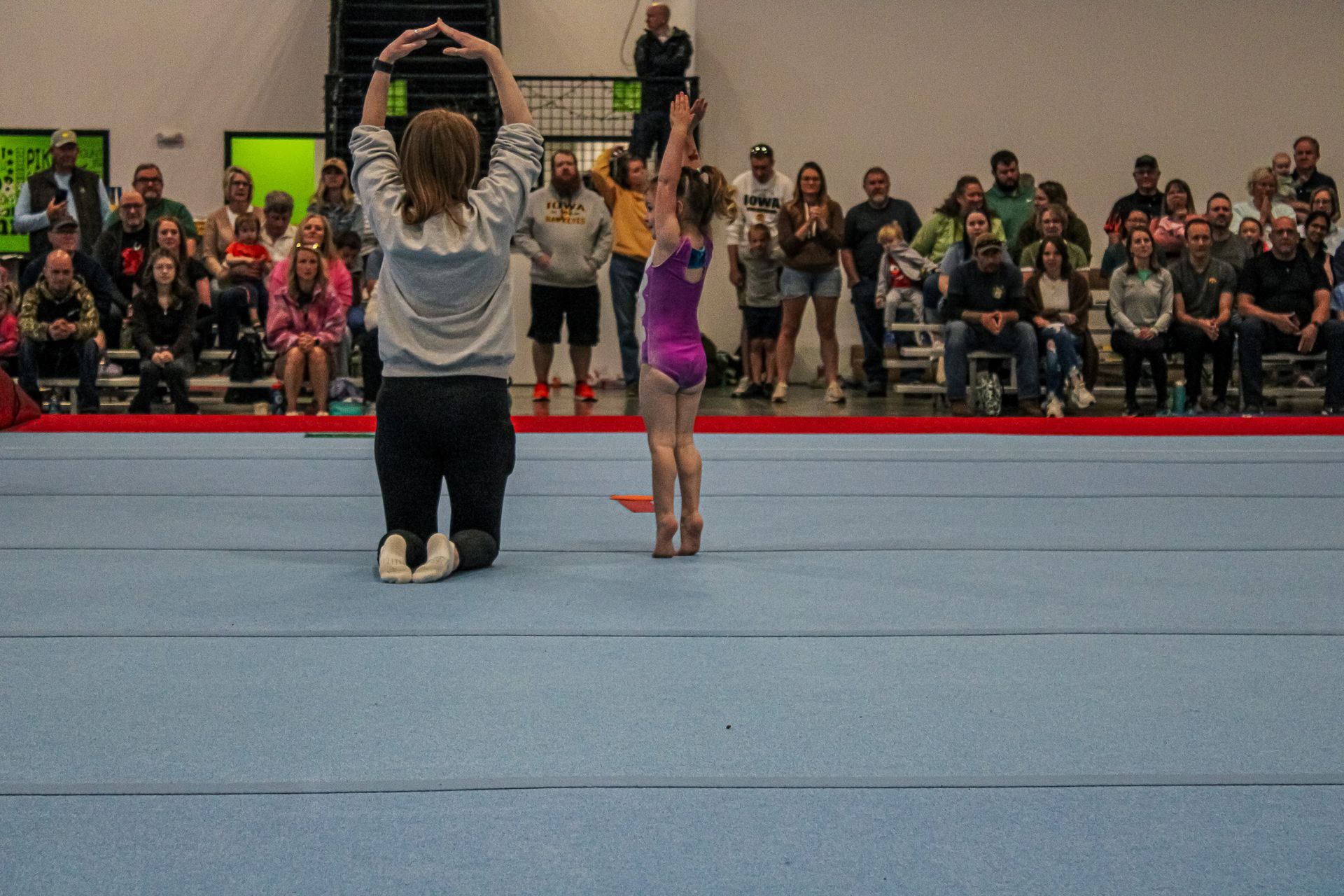 Coach kneeling before a child gymnast on a blue mat in a gym, with spectators watching in the background
