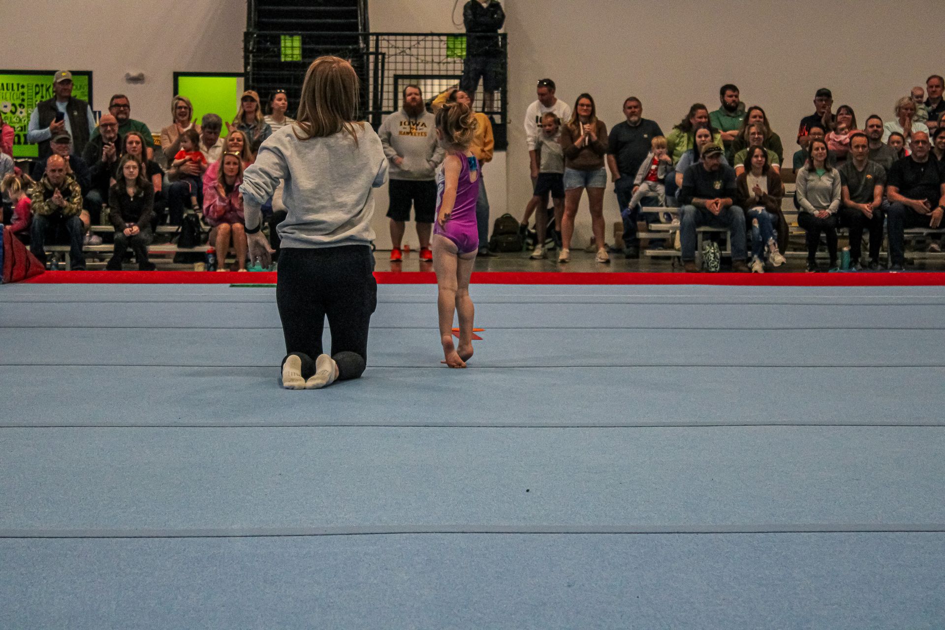 Gymnastics floor routine with a coach standing beside a young athlete in a gymnasium, spectators in the background