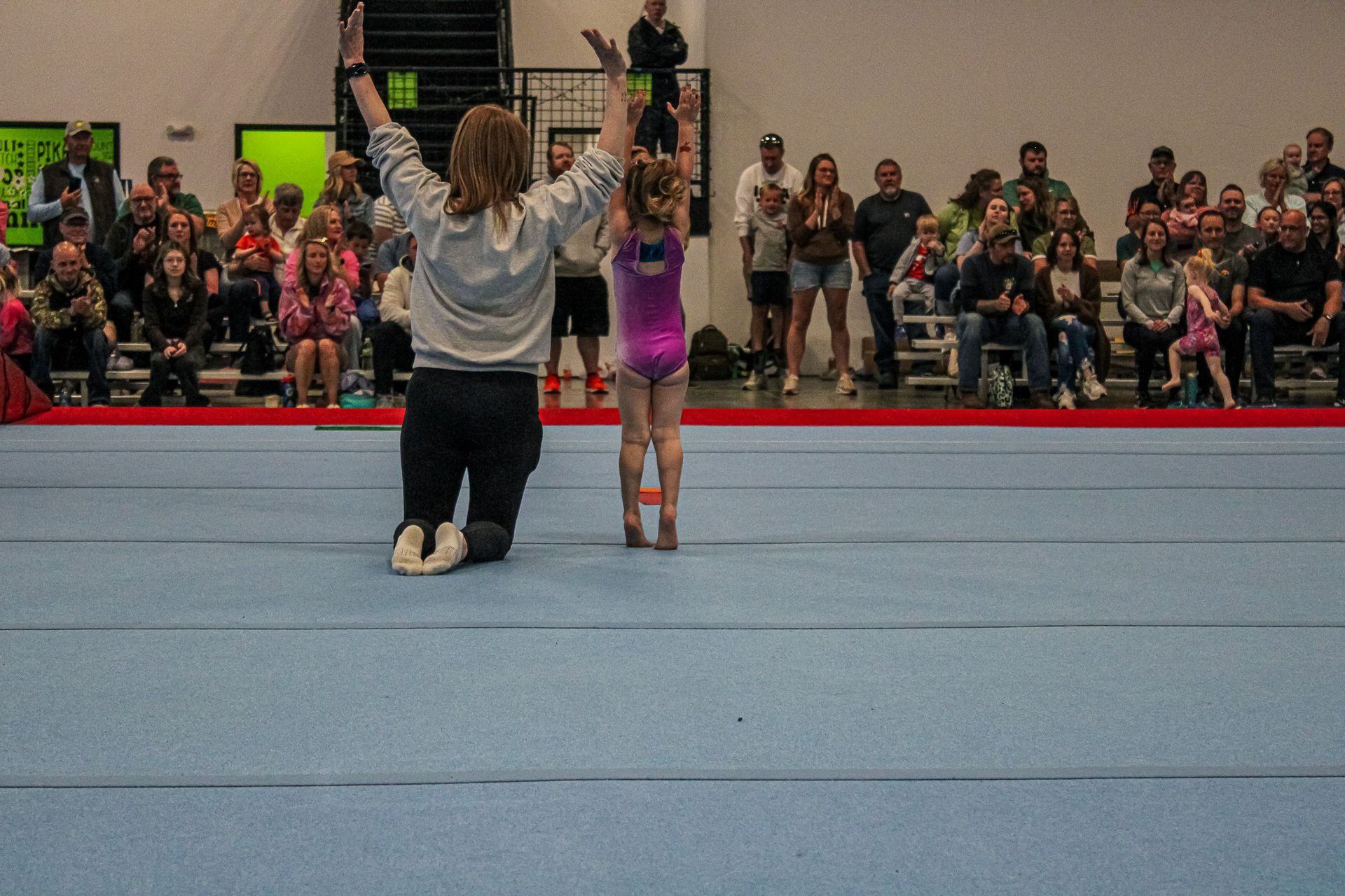 Two gymnasts perform a floor routine, one kneeling with arms raised and one standing on shoulders, before an audience.