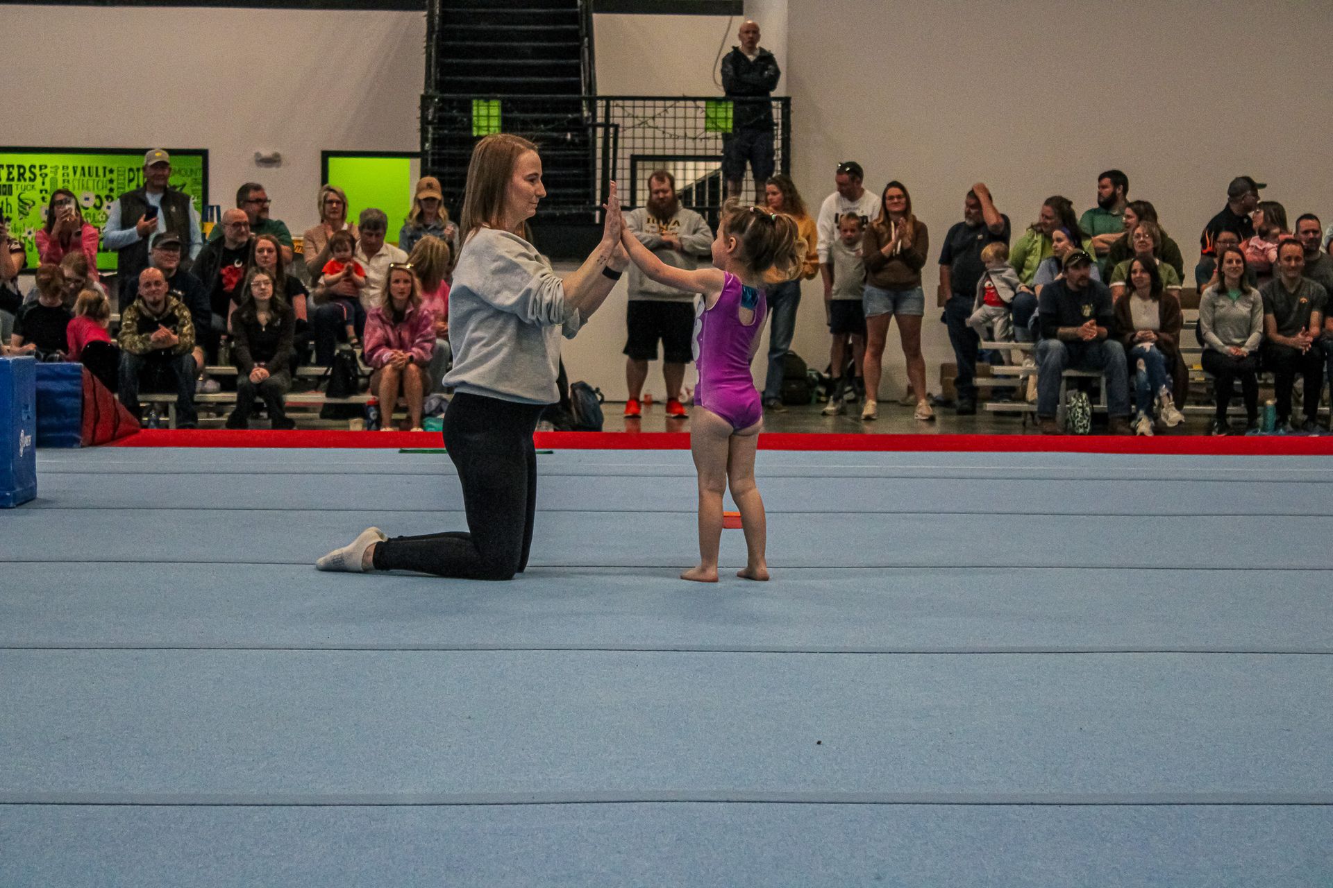 Gymnast kneels on floor while a young child balances on her hands in an indoor gymnasium