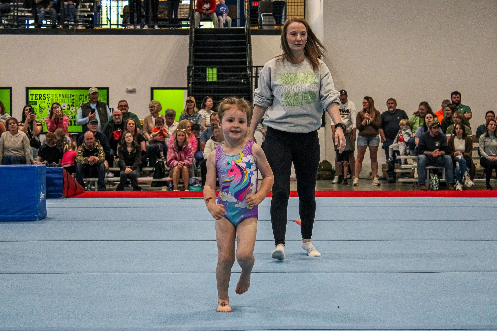 Young gymnast in colorful leotard walking on a blue floor with a coach behind her in an indoor gymnasium