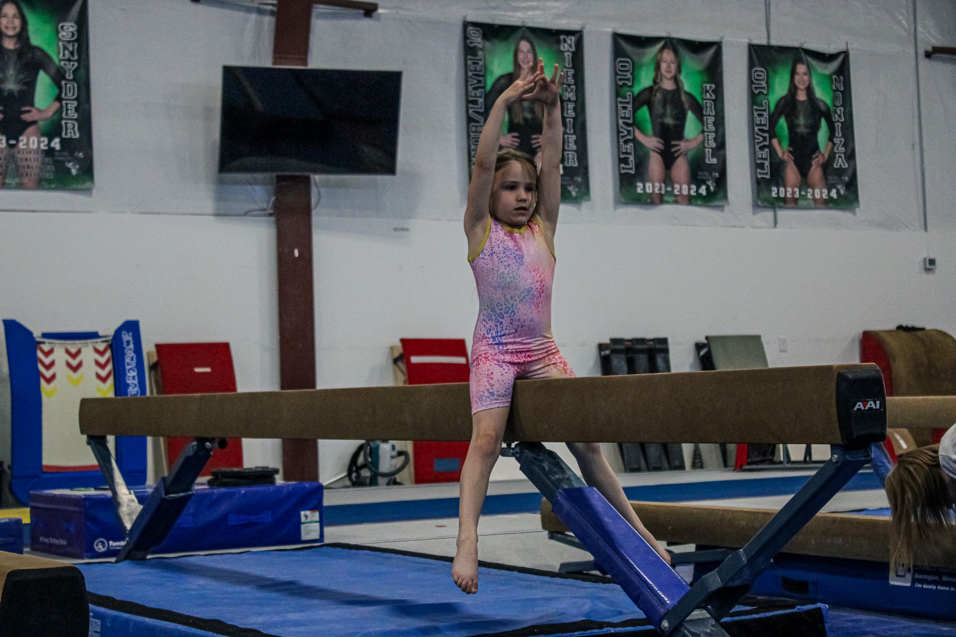 Young gymnast in pink leotard balancing on a beam in a gym, arms raised overhead
