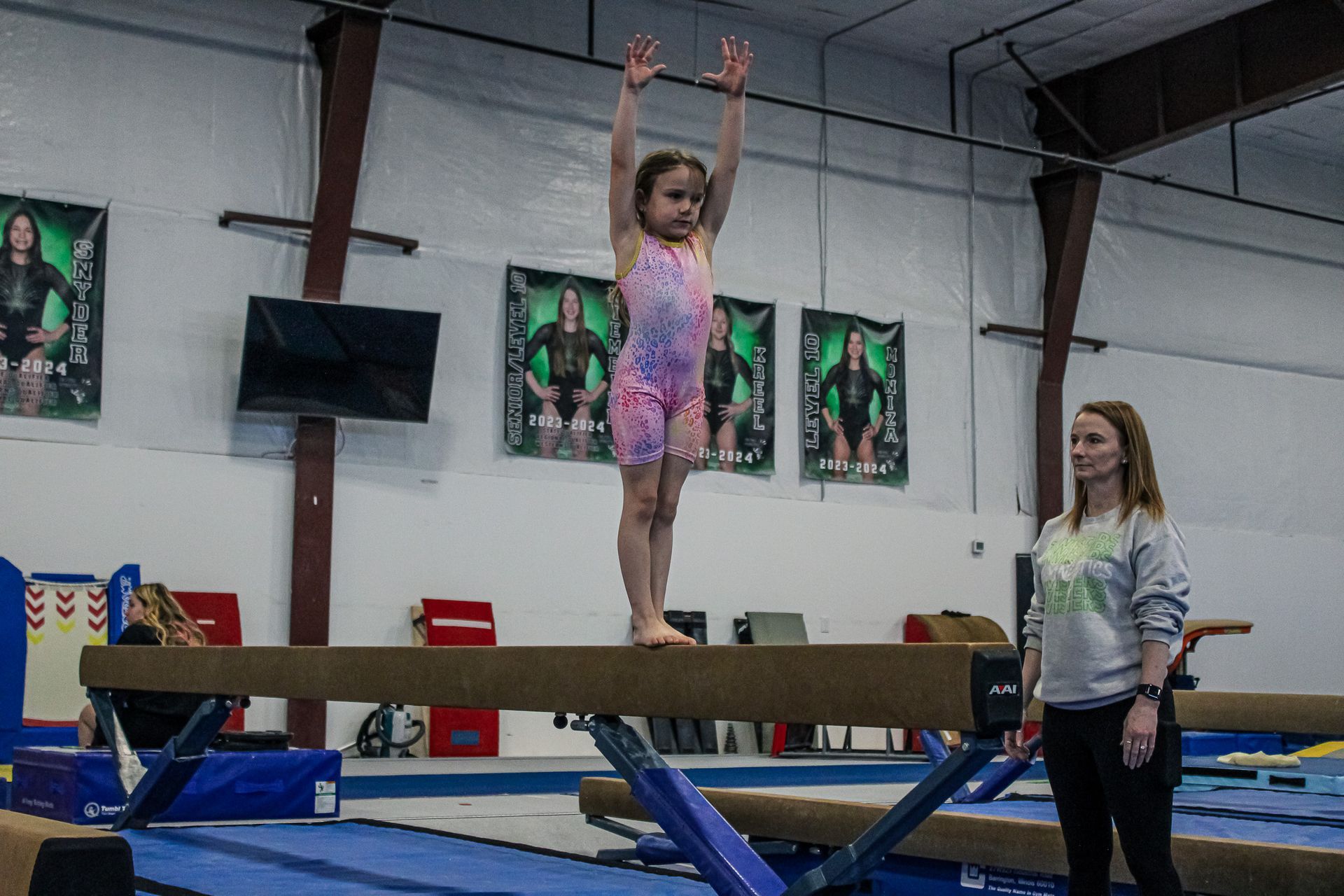 Gymnast balancing on a beam with arms raised in an indoor gym while a coach watches nearby