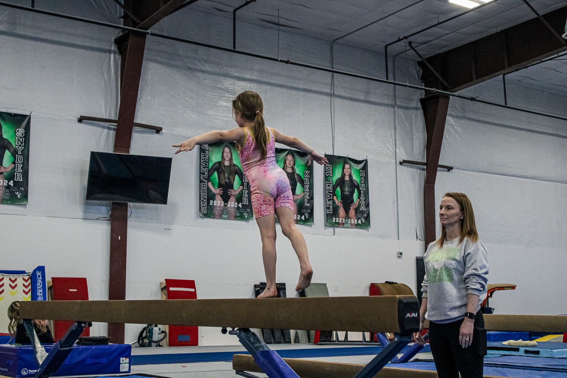 Gymnast balancing on a beam in a gym while a coach watches nearby.