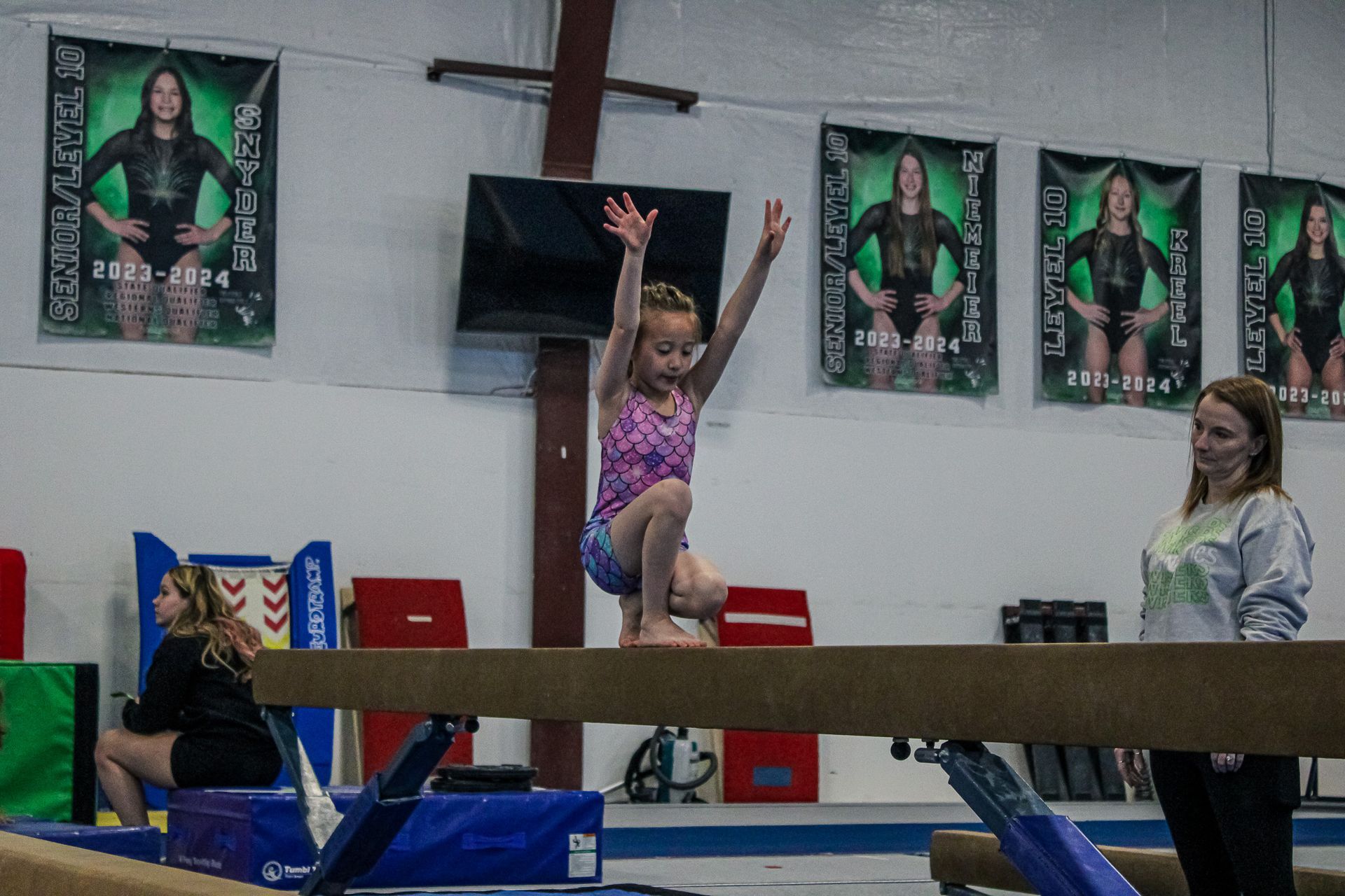 Gymnast balancing on a beam with arms raised, coaches spotting in a gymnasium with posters on the wall.