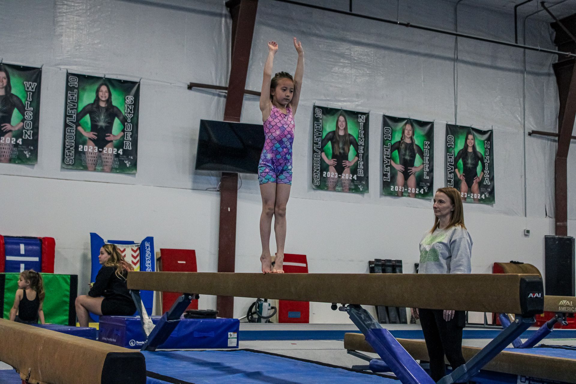 Gymnast balancing on a beam with arms raised in an indoor gym, with a coach nearby.