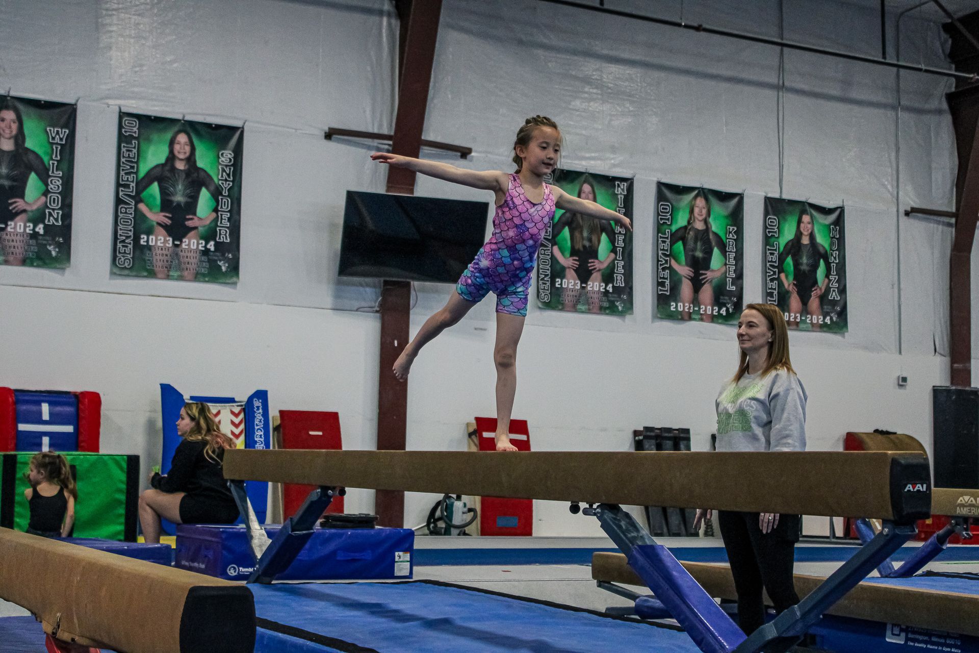 Young gymnast balancing on a beam in a gym, with coaches watching nearby