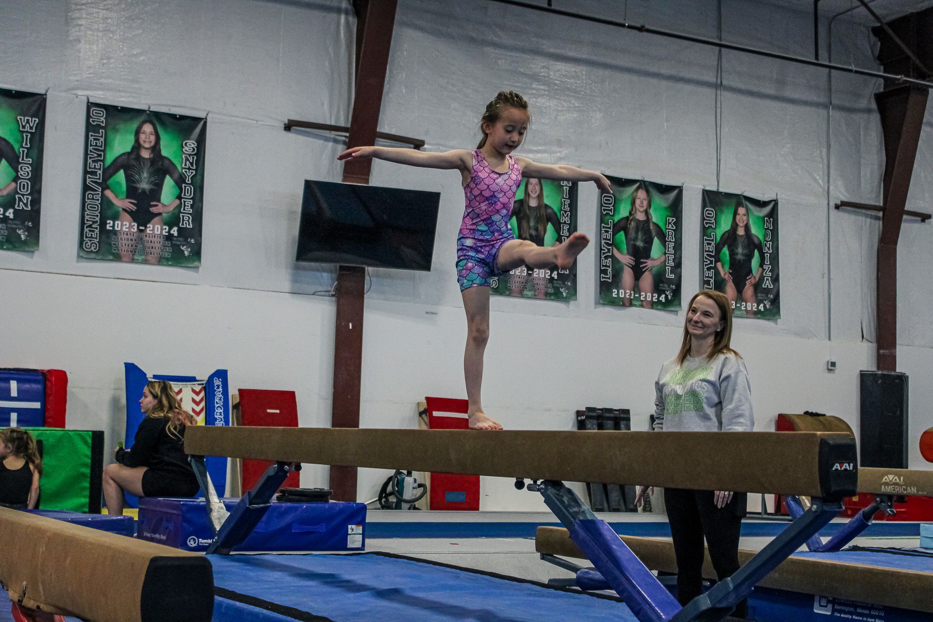 Gymnast balancing on a beam in a gym, with a coach watching nearby.