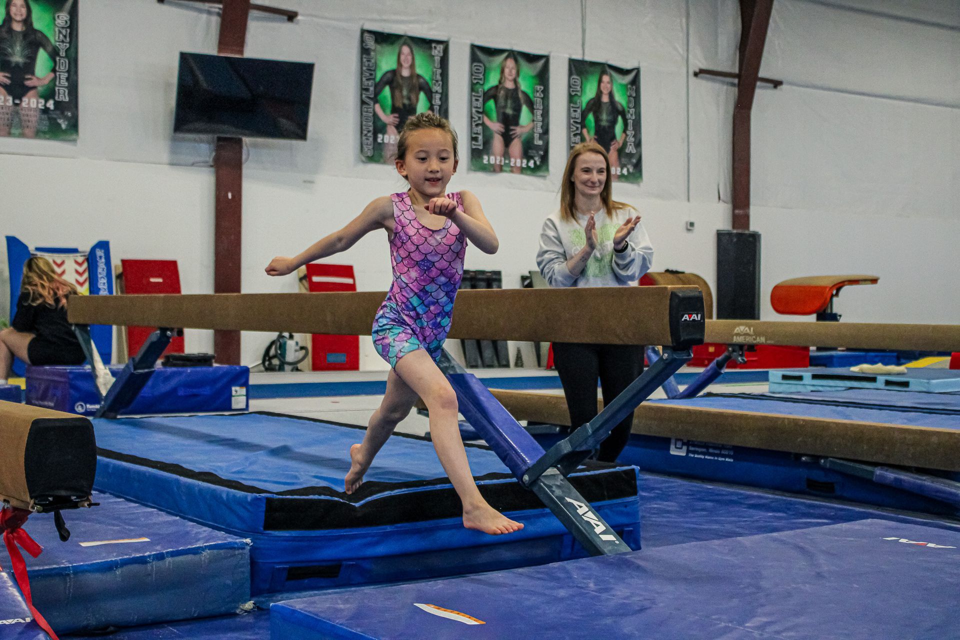 Child balancing on a gymnastics beam with a coach in a gymnasium, blue mats below