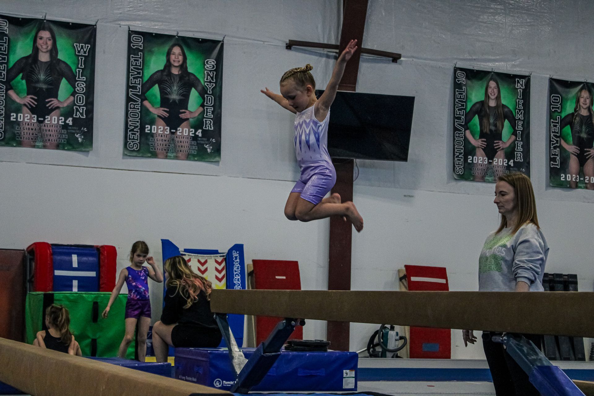 Gymnast mid-air on a balance beam in a gym, with posters and mats in the background.