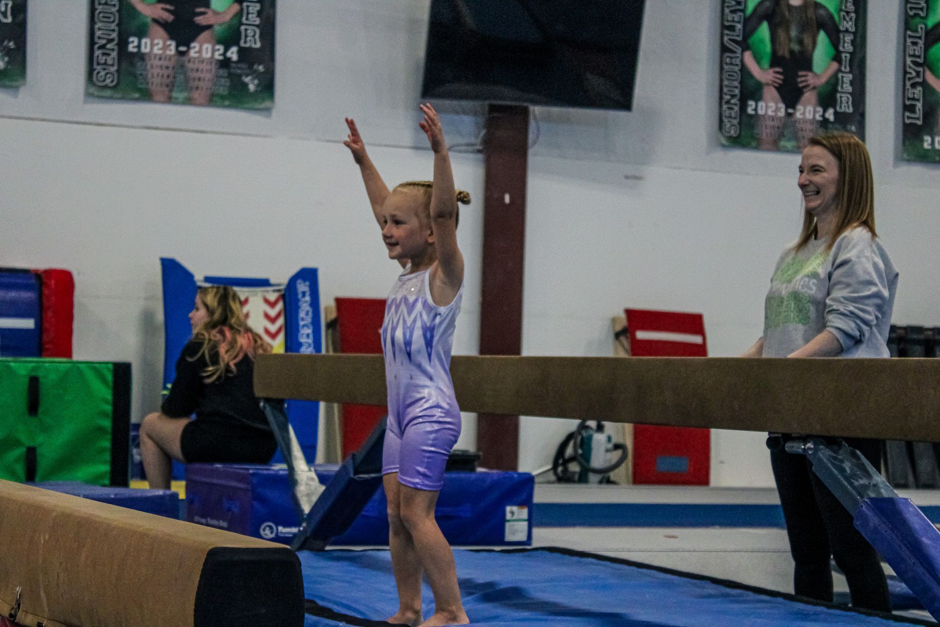 Gymnast balancing on a beam with arms raised, coach standing nearby in an indoor gym.