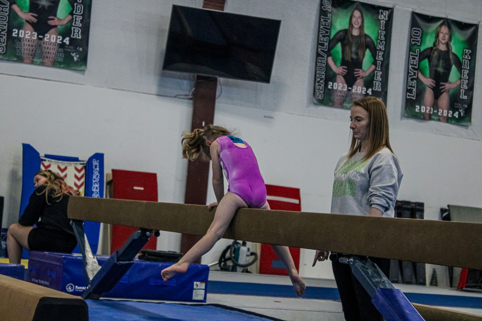 Gymnast balancing on a beam in a gym while a coach watches nearby