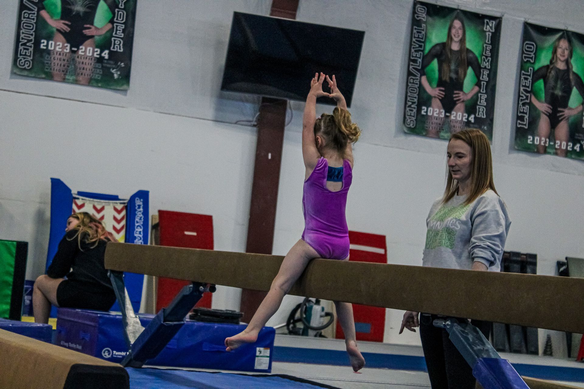 Gymnast balancing on a beam with arms raised, in a gym with coaches and posters in the background.