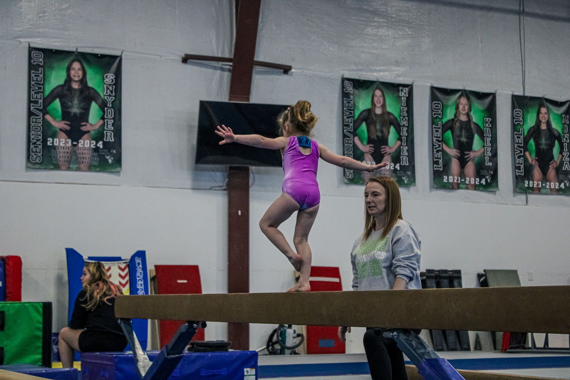 Gymnast balancing on a beam with a coach spotting her in an indoor gym