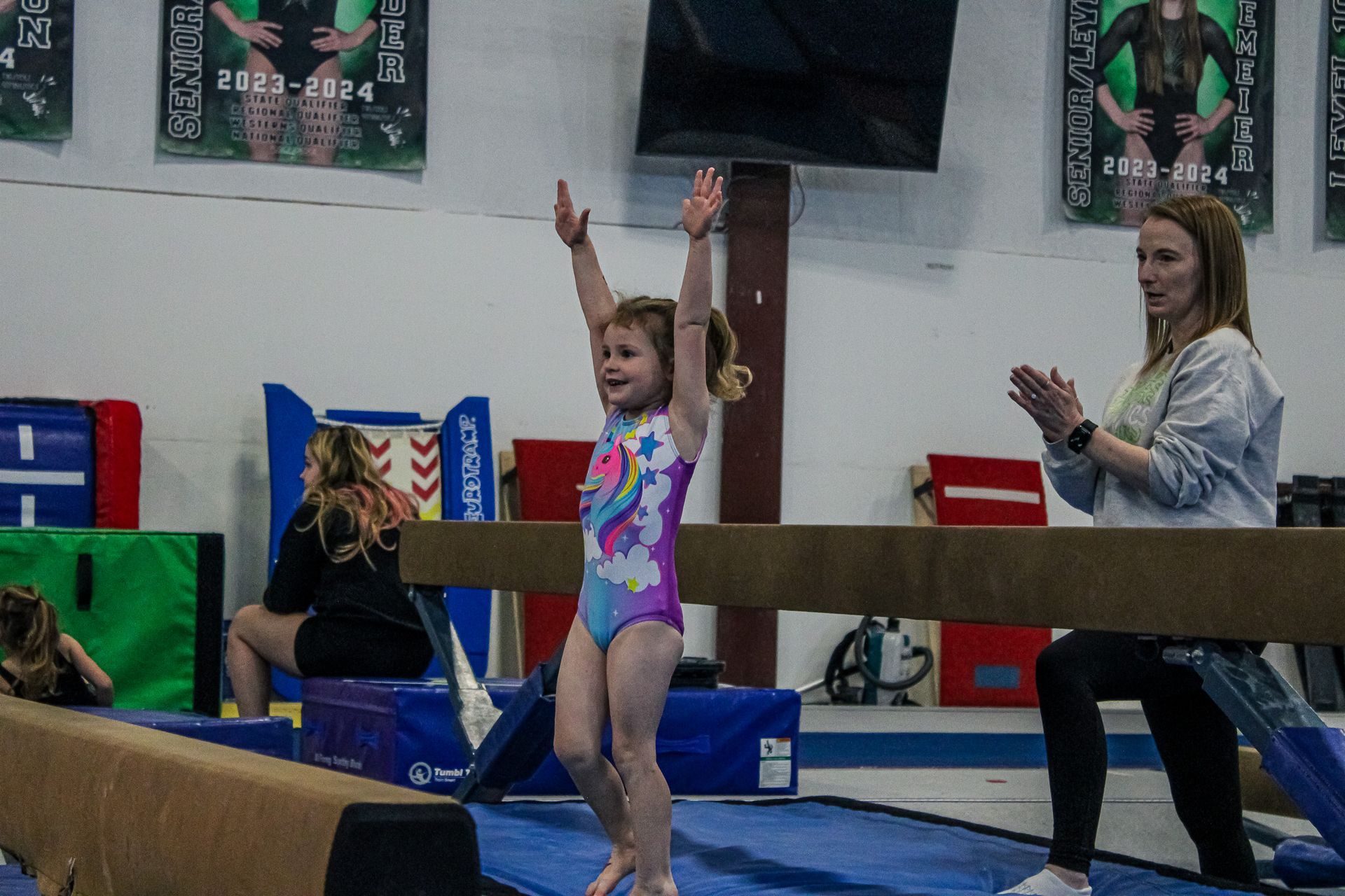 Young gymnast in a colorful leotard raises her arms on a balance beam while a coach applauds nearby.
