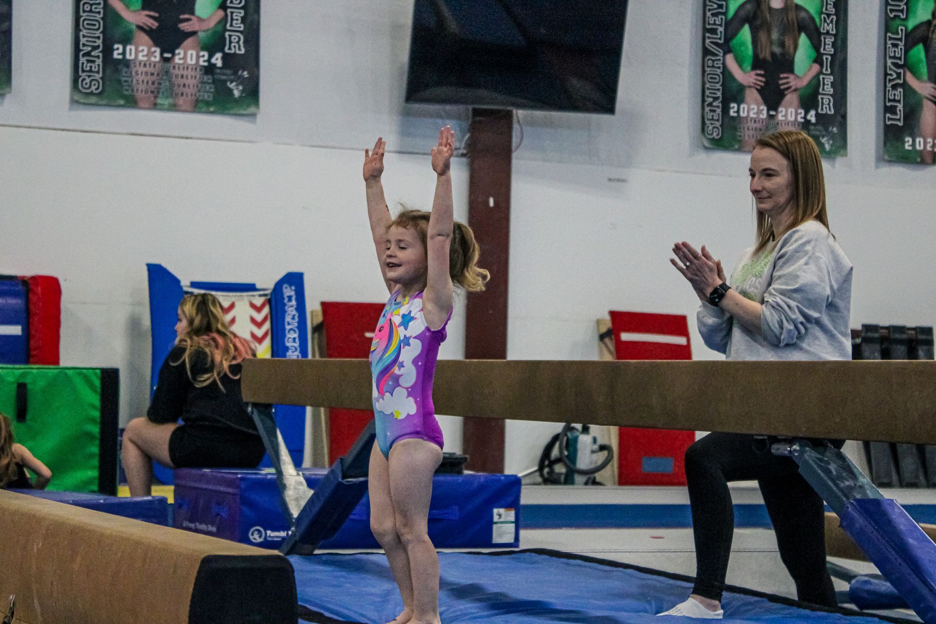 Gymnast balancing on a beam in a gym, arms raised, with a coach watching nearby