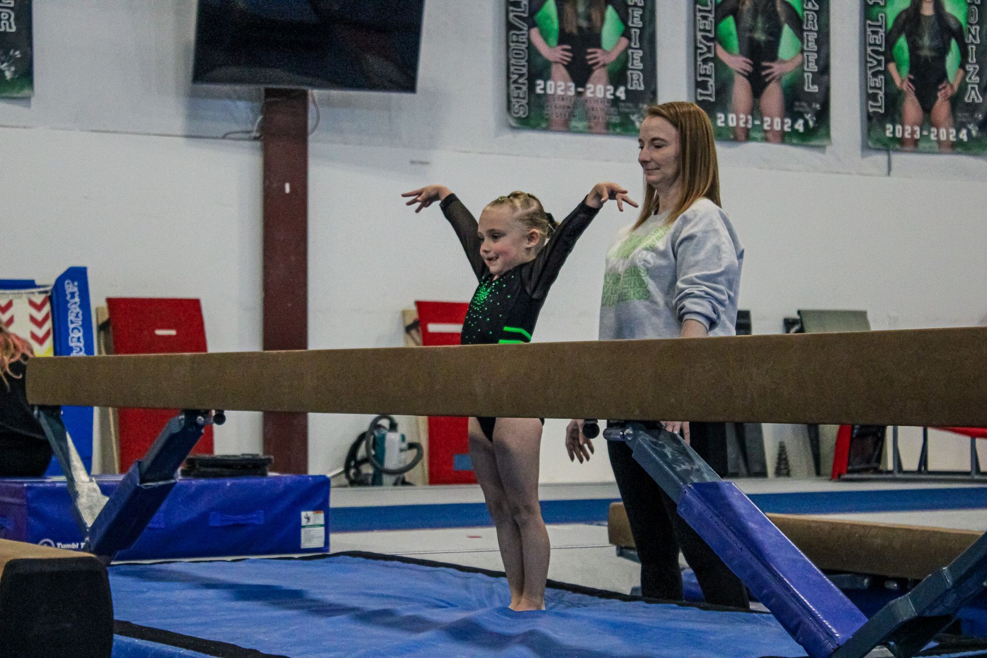 Gymnast balancing on a beam with a coach spotting on a blue mat in a gym