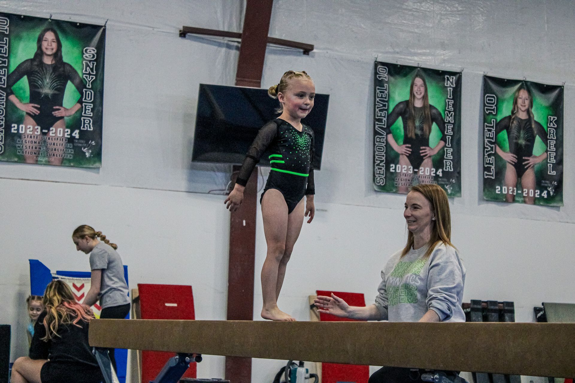 Young gymnast standing on a balance beam while a coach watches in an indoor gym.