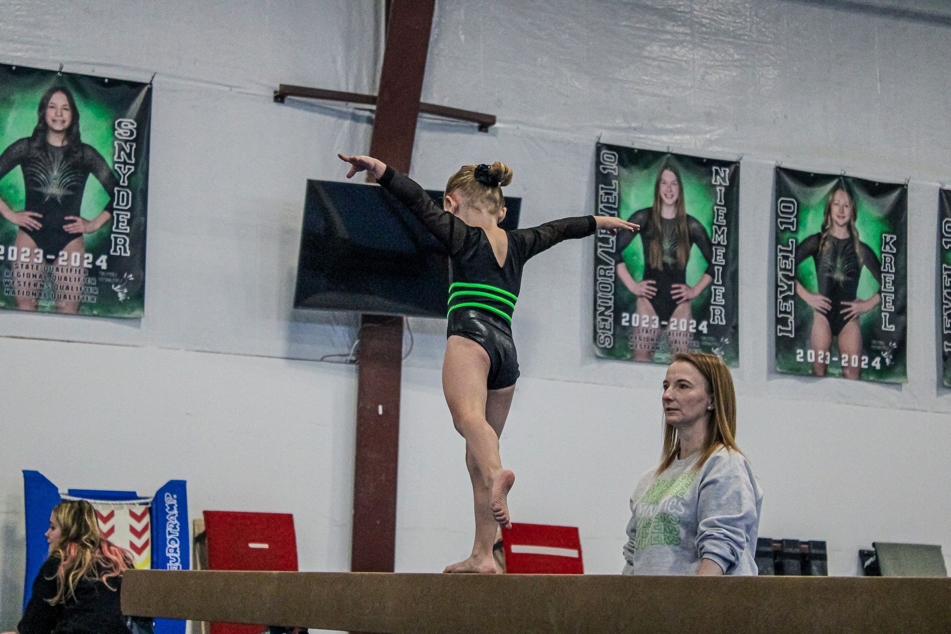 Gymnast balancing on a beam while a coach watches nearby under posters on the wall.
