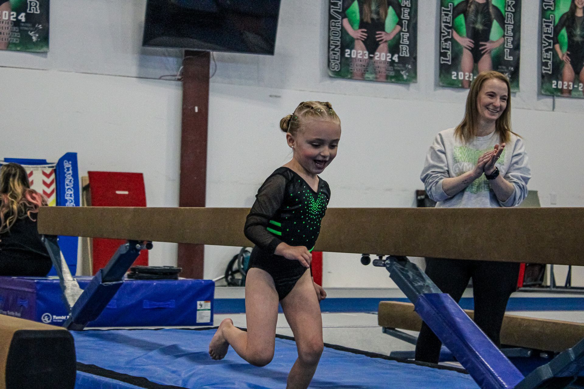 Young gymnast in a sparkly black leotard kneels on a balance beam in a gym, with a coach clapping nearby.