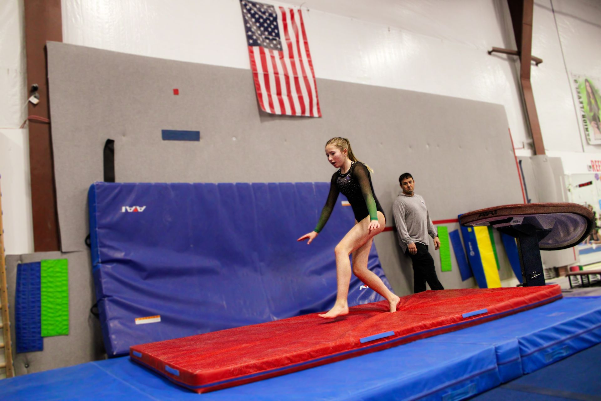 Gymnast balancing on a red mat in a gym, with a coach nearby and an American flag on the wall.