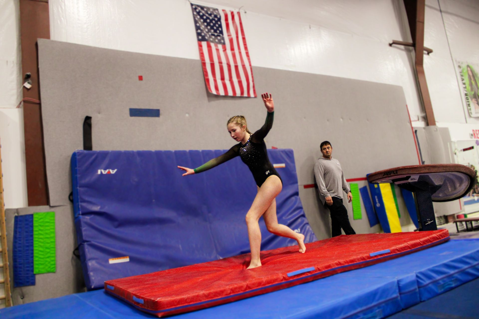 Gymnast balancing on a red mat in a gym, with an American flag and coach nearby