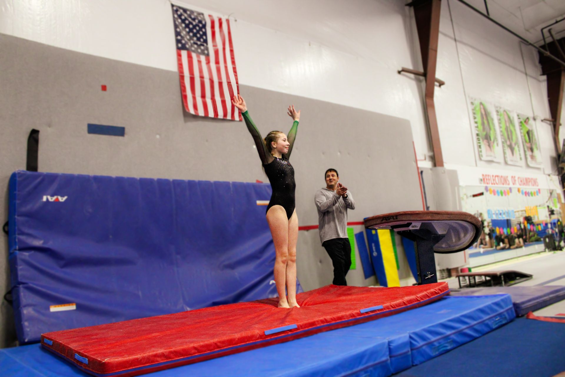 Gymnast on a red mat with arms raised, beside a coach in a gym.