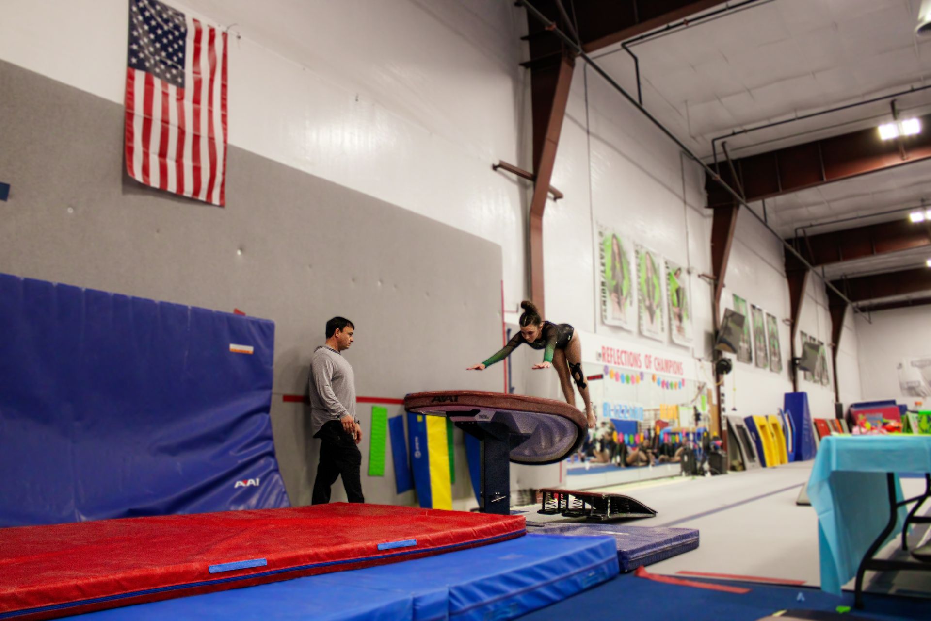 Gymnastics gym with a person on a vault and another standing nearby, mats and an American flag in the background