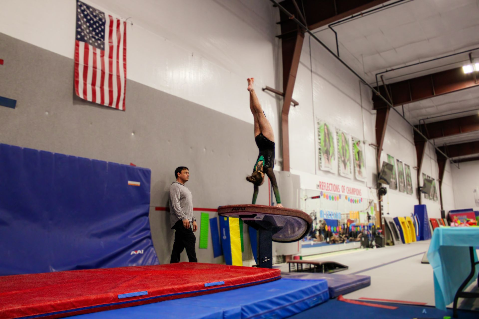 Gymnast balanced upside down on a bar in a gym, with a coach watching nearby.