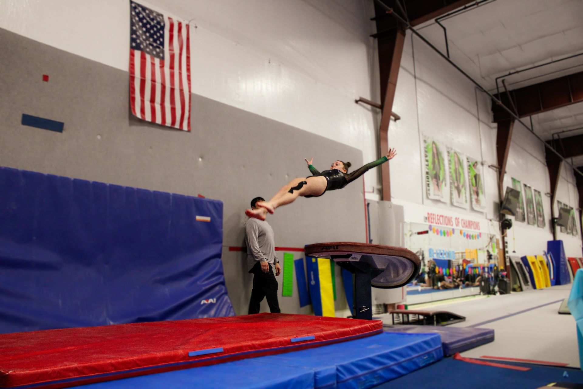 Gymnast flipping over a vault in an indoor training gym with a coach nearby and mats below