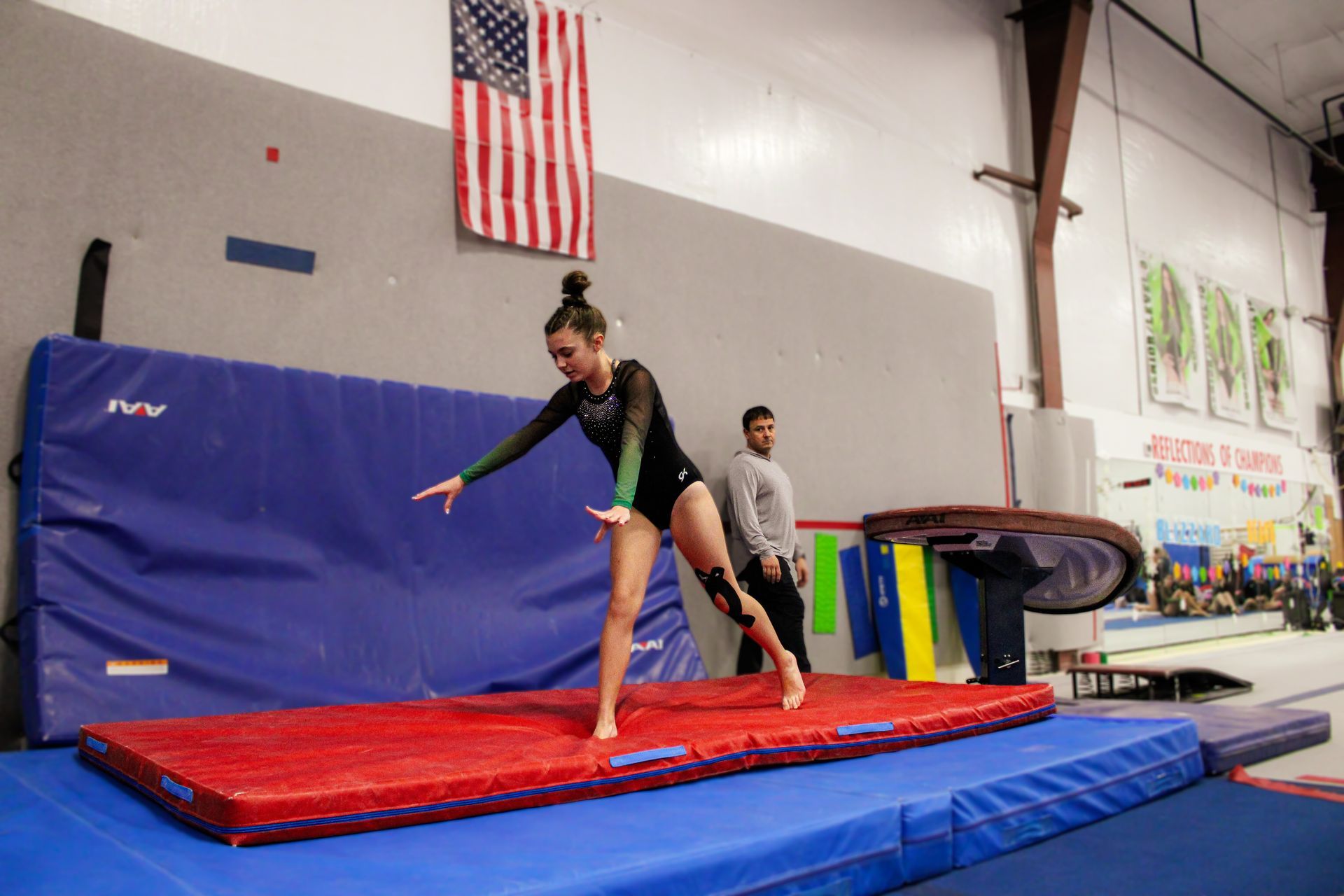 Gymnast balancing on a red mat in a gym, with a coach nearby and an American flag overhead