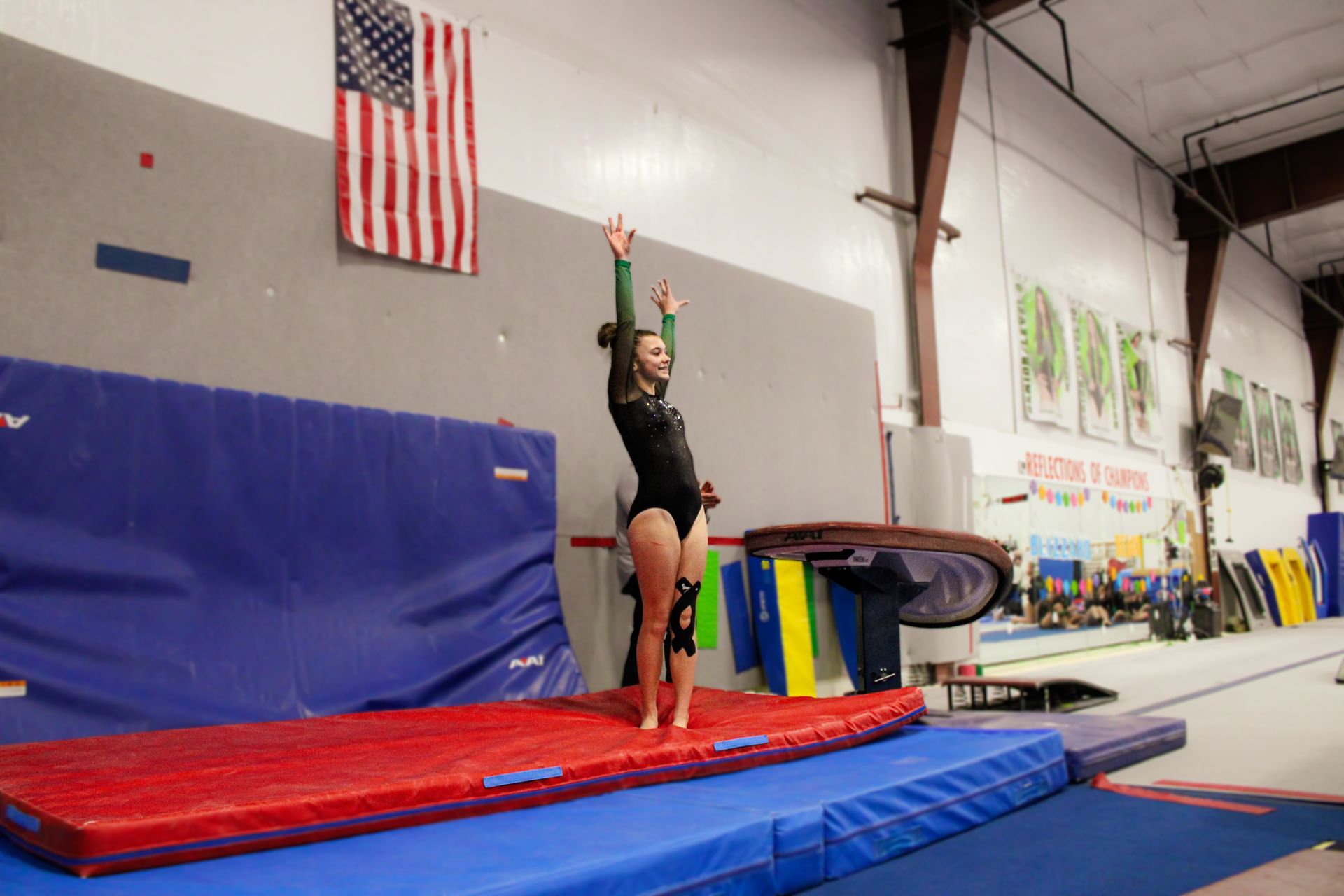 Gymnast balancing on a red mat in a gym, arm raised beside a balance beam.