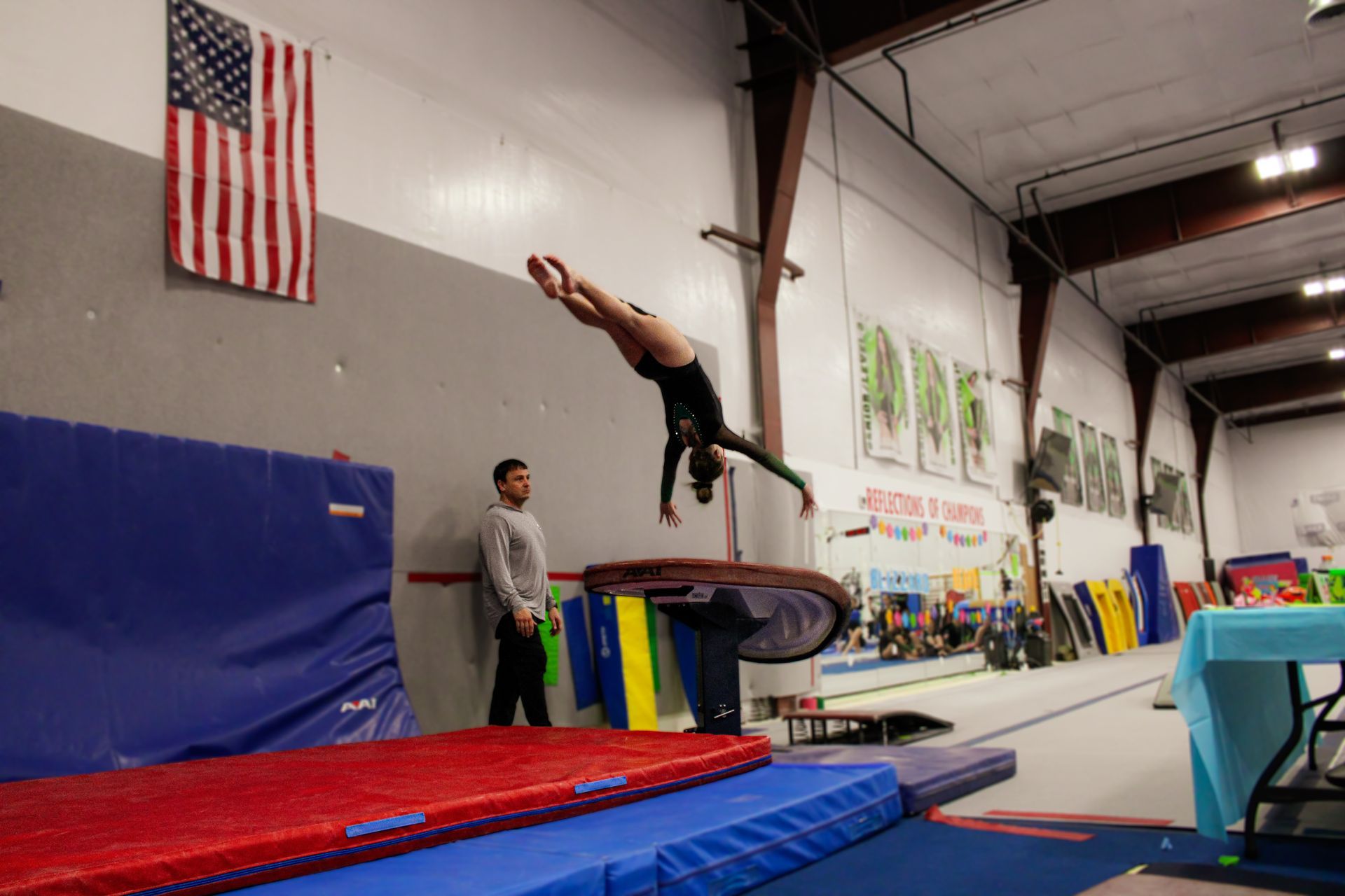 Gymnast midair on a springboard in a gym, with blue and red mats and an American flag in the background