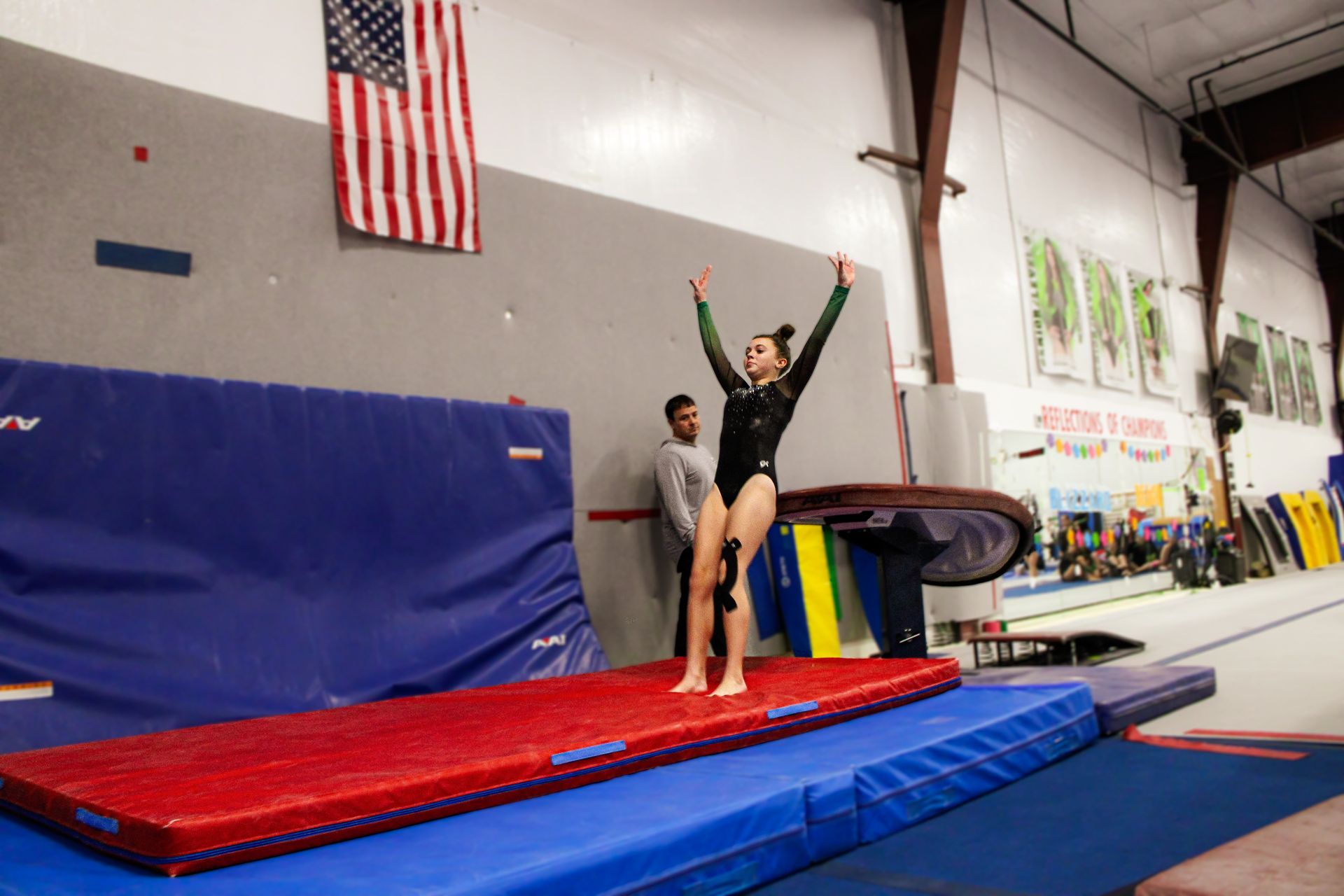Gymnast balancing on a beam in a gym, arms raised on a red mat beside blue pads.