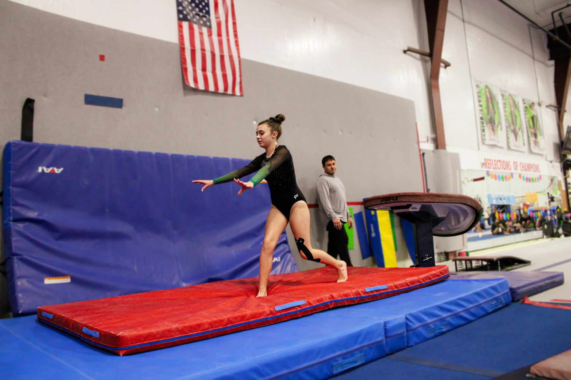 Gymnast balancing on a red mat in a gym with blue padding and an American flag overhead