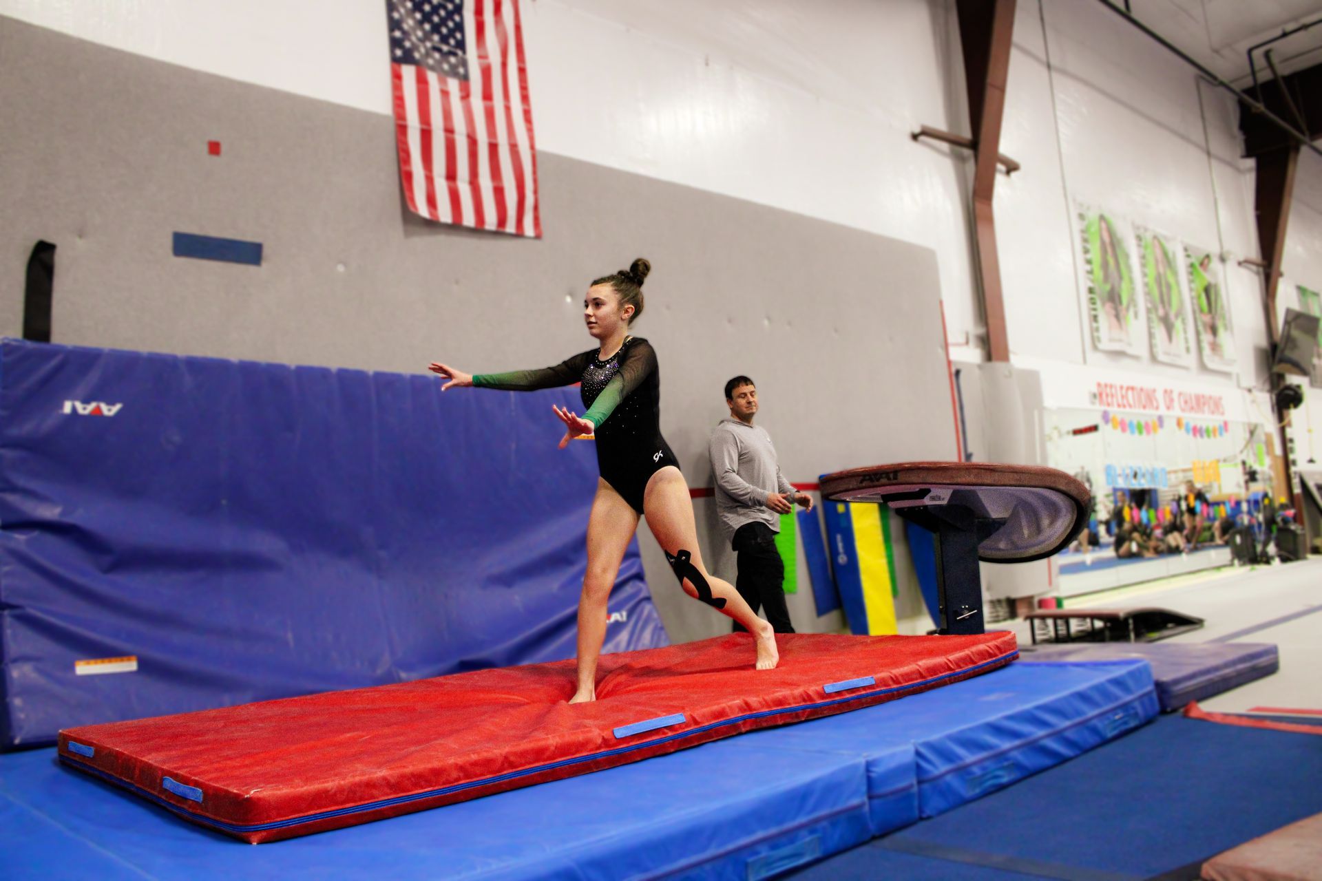 Gymnast balancing on a red mat in a gym, with a coach nearby and an American flag on the wall.