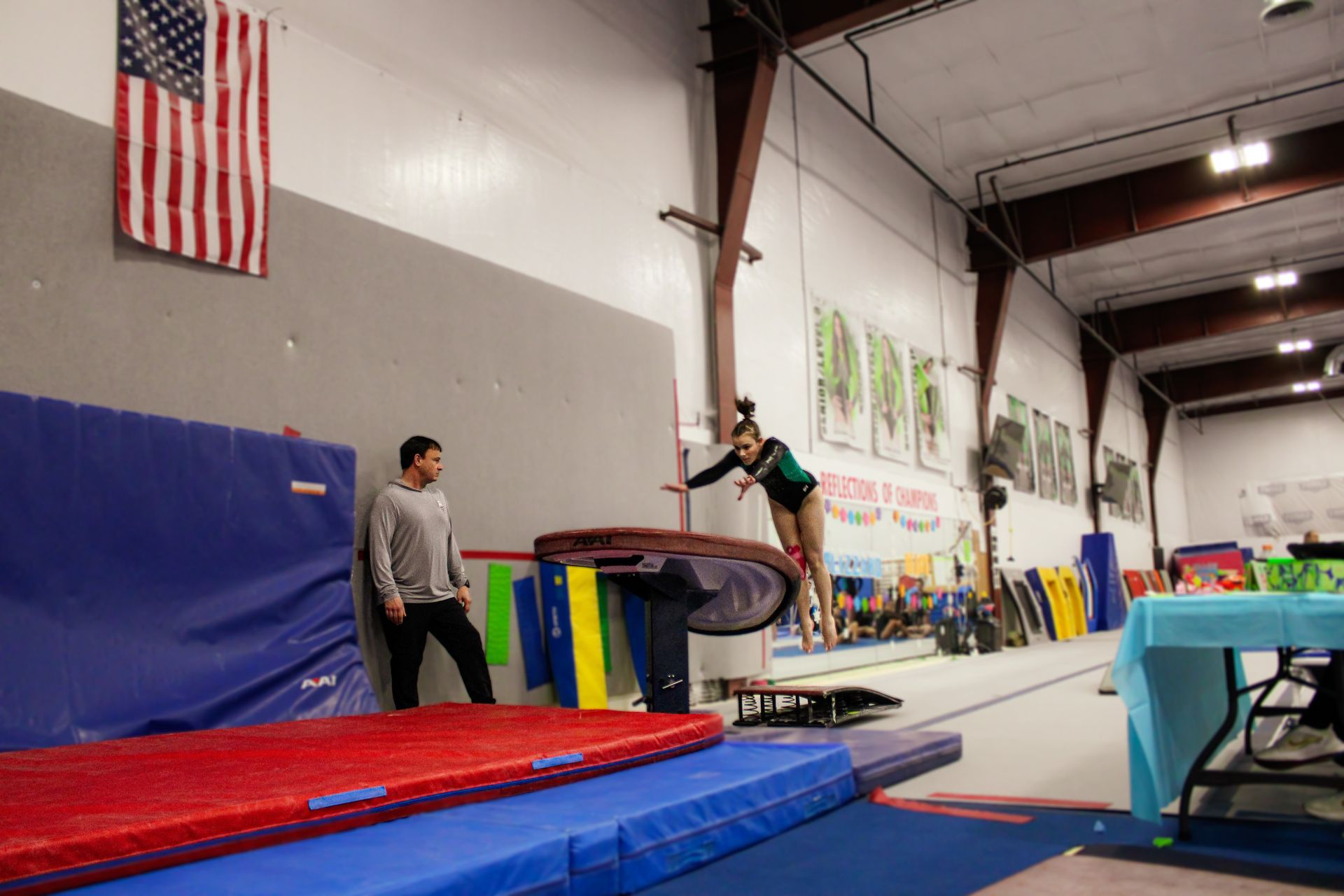 Gymnast mid-air on trampoline in an indoor training gym, with a coach and mats nearby.