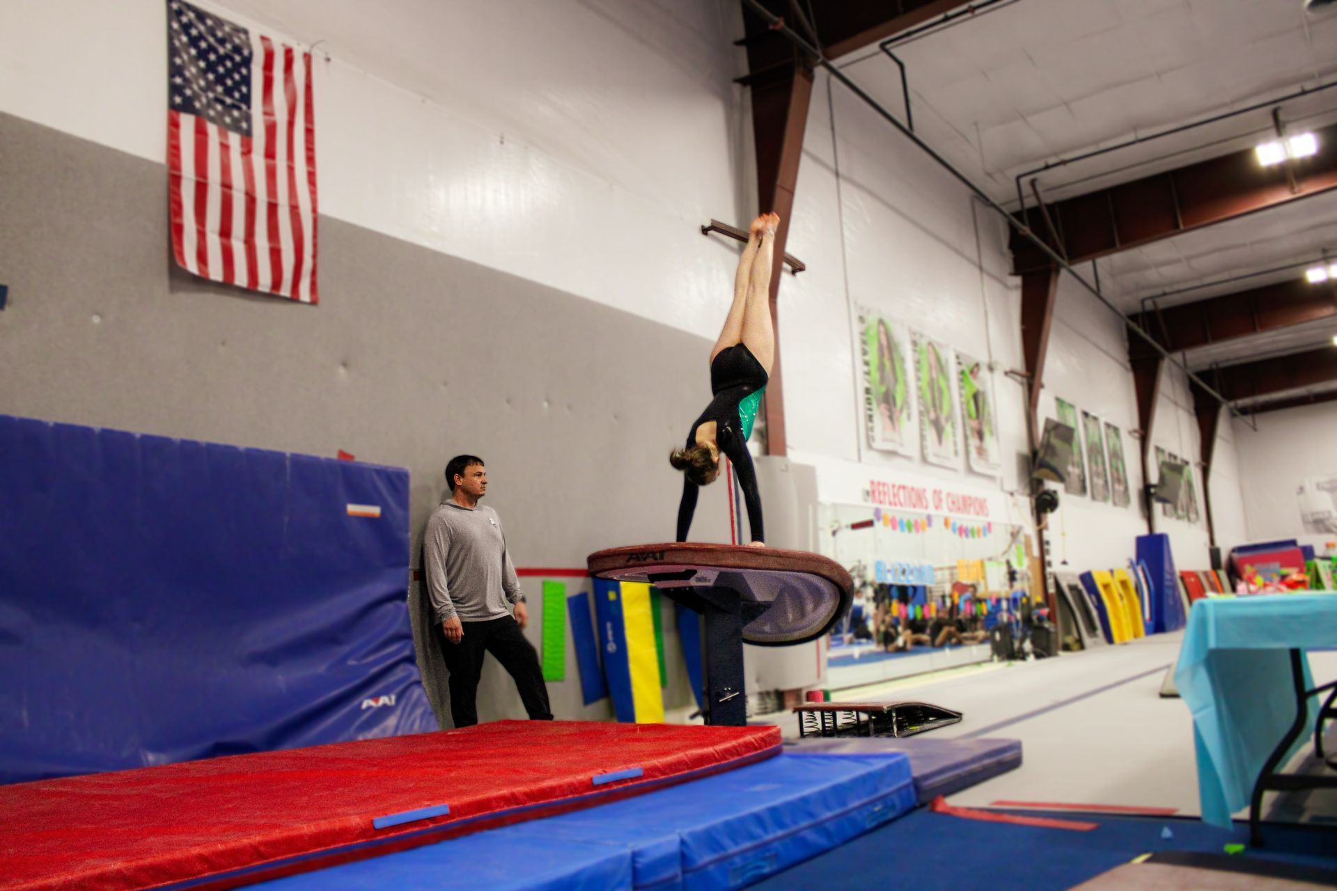 Gymnast balancing on a beam in a tumbling gym, with mats and a coach nearby