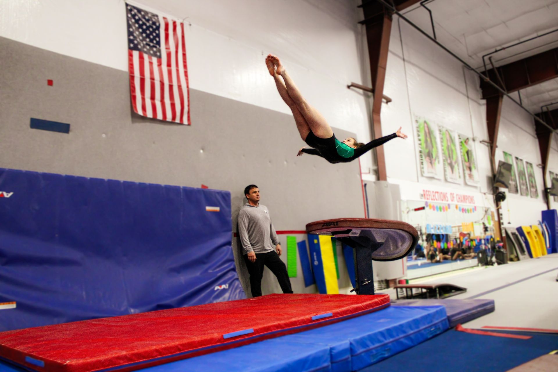 Gymnast flipping over a vault in a gym with blue and red mats and an American flag overhead
