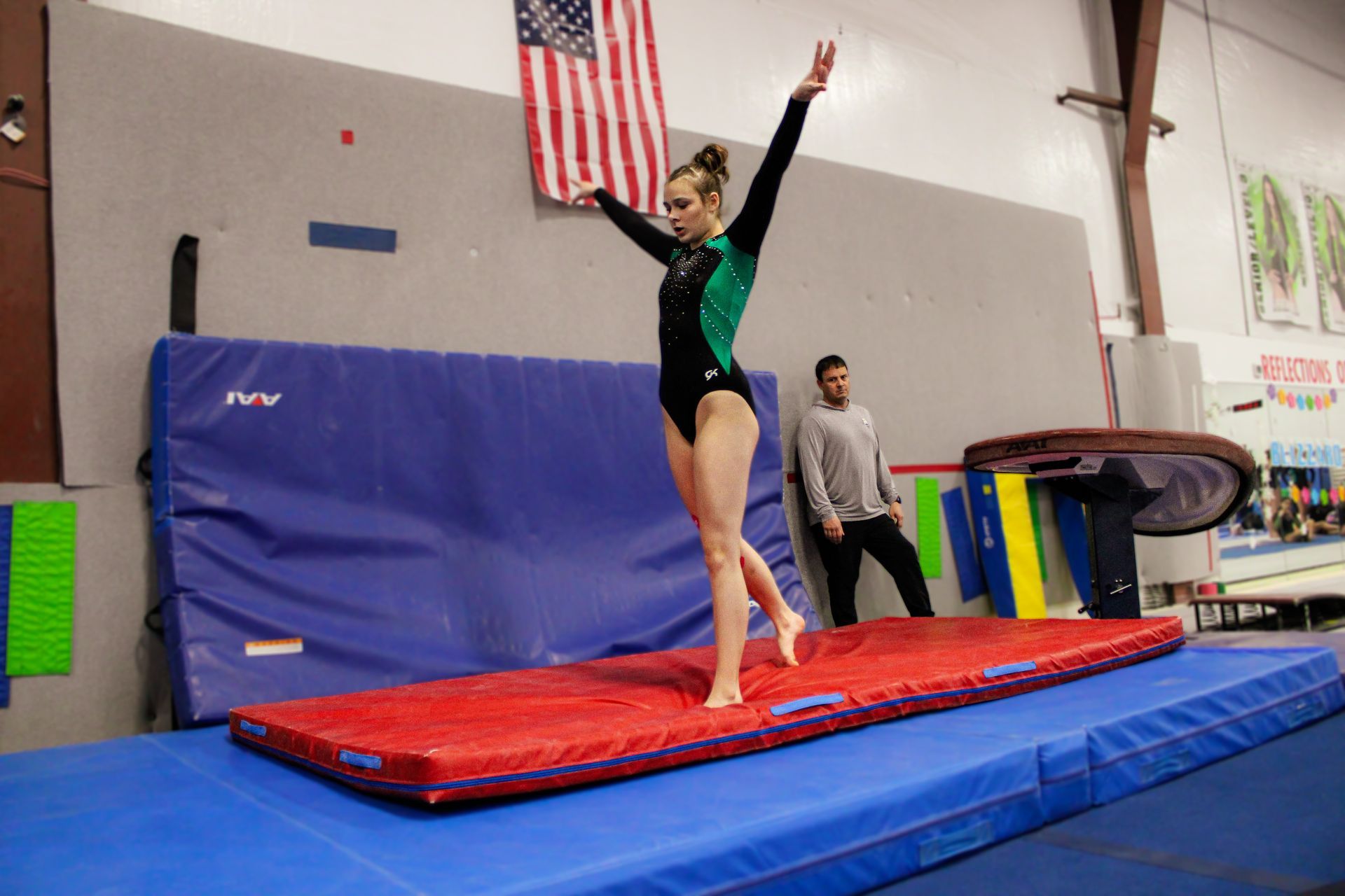 Gymnast balancing on a red mat in a gym, arms raised, with a coach nearby and an American flag in the background