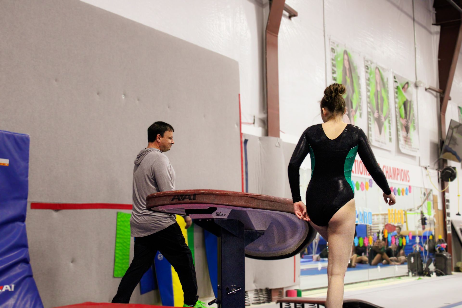 Gymnast in black leotard walks toward a coach beside a balance beam in a gym.