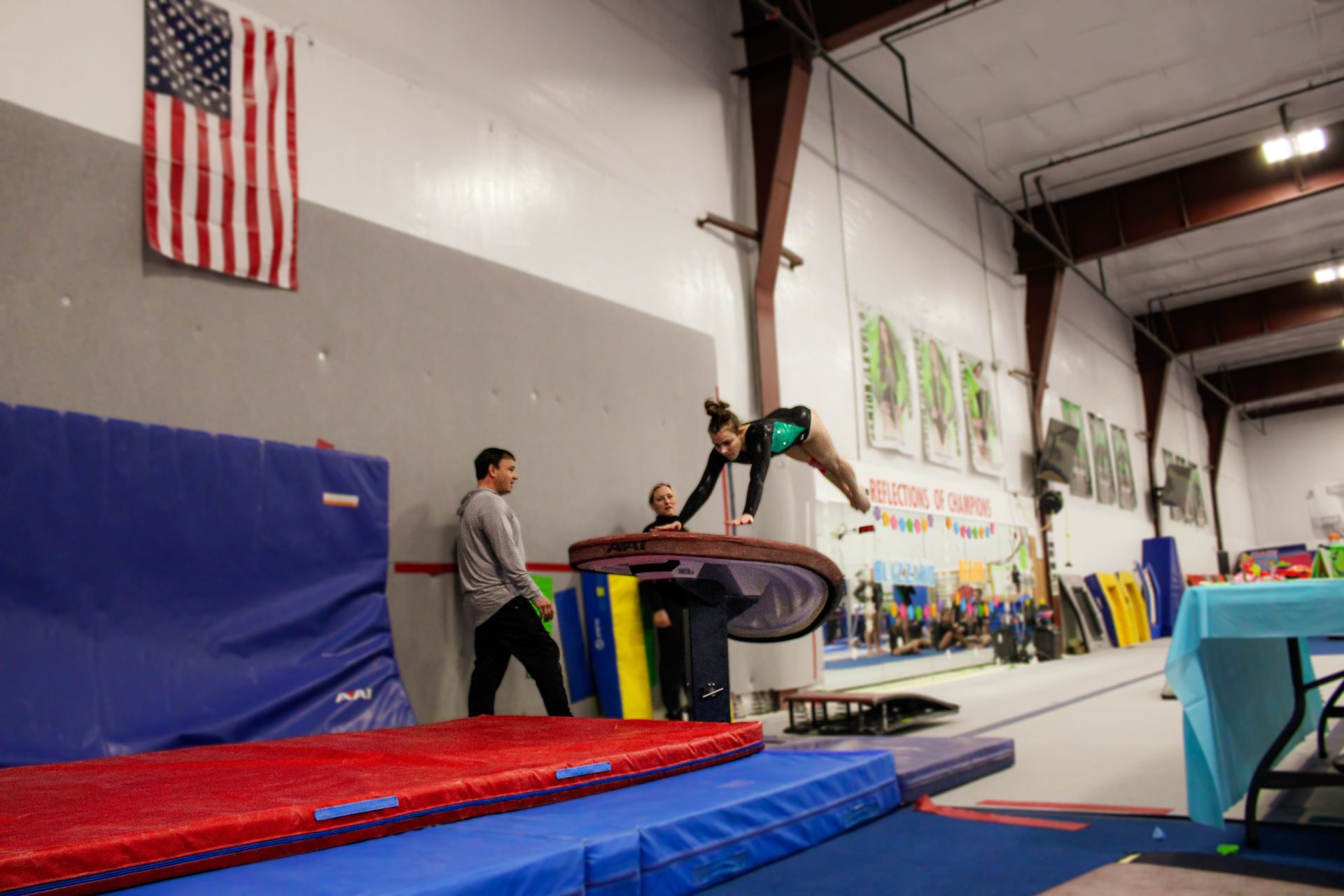 Gymnast balancing on a vault box in a gymnastics gym, with coach nearby and red-blue mats below