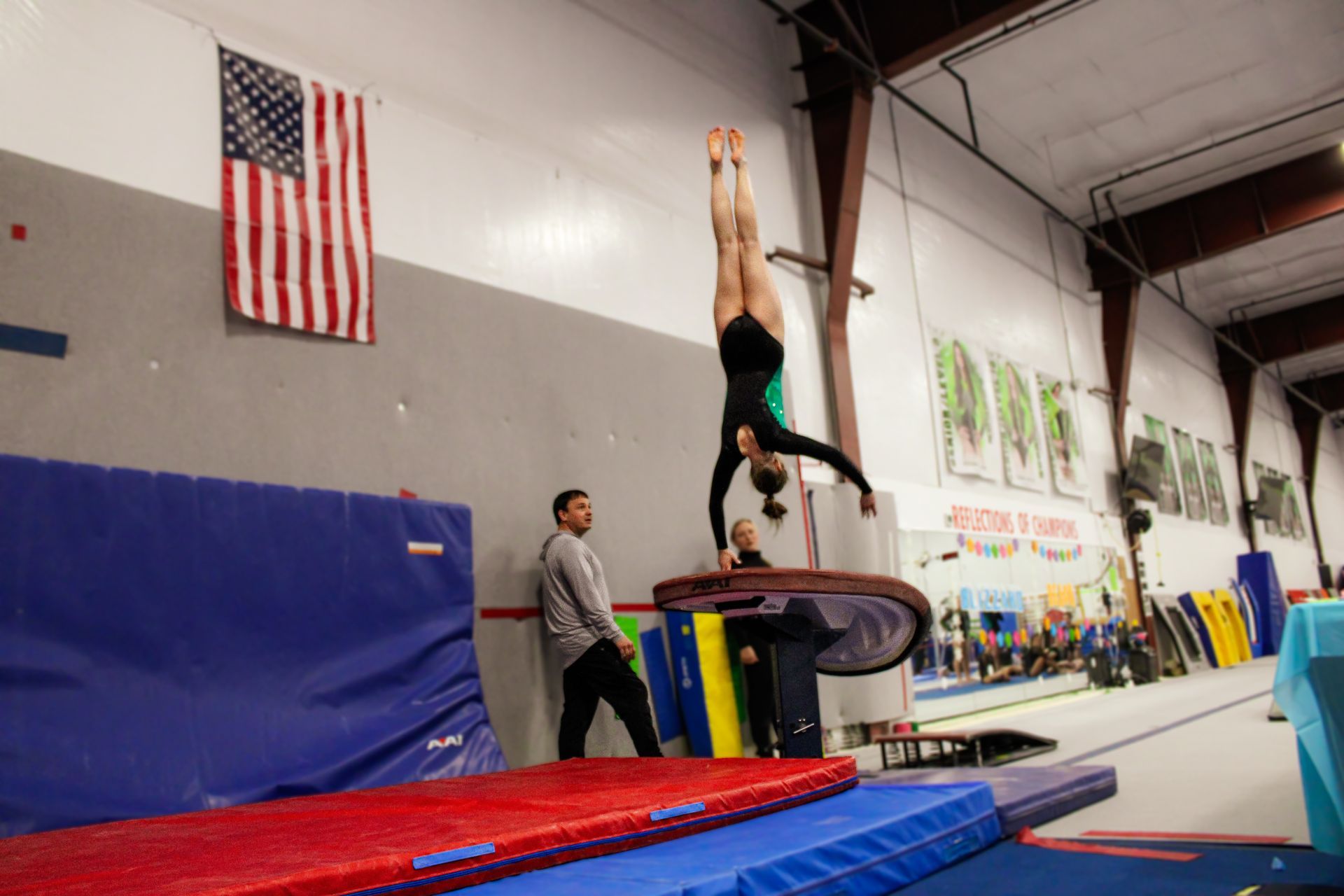 Gymnast doing a handstand on a vault in an indoor training gym, with a coach nearby.