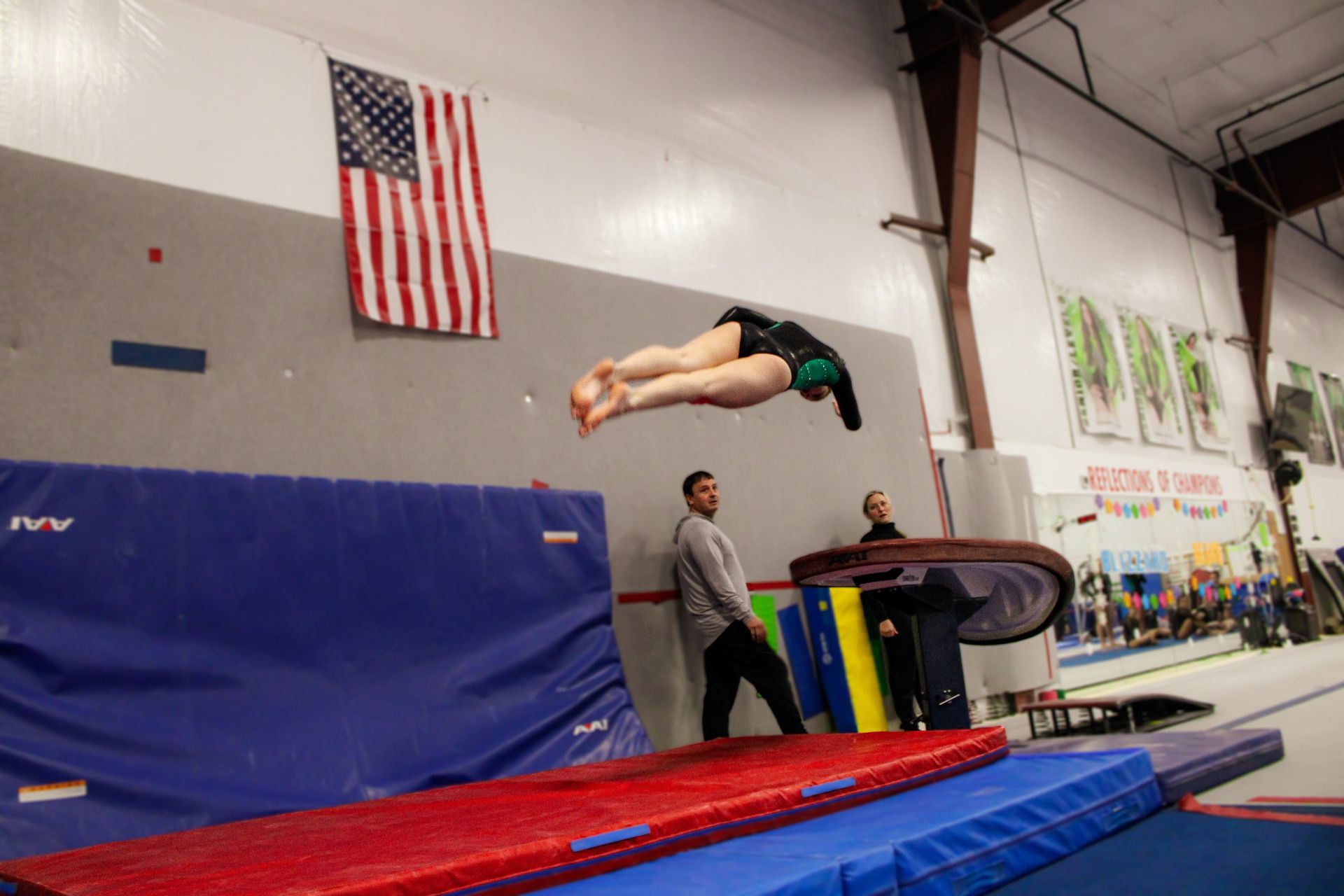Gymnast midair over a vault in a gym with a U.S. flag on the wall