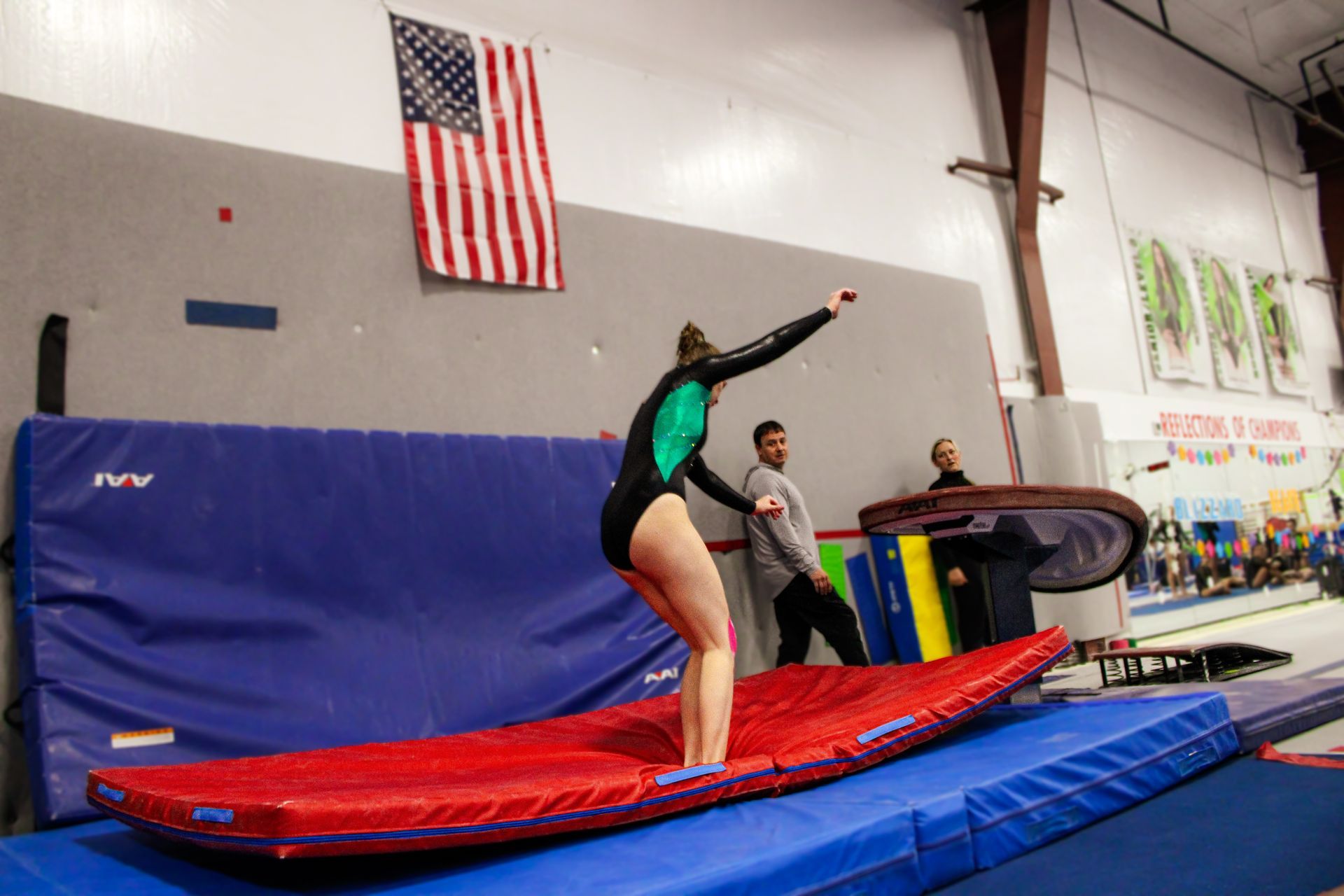 Gymnast balancing on a red mat in an indoor gym, with a coach and American flag in the background